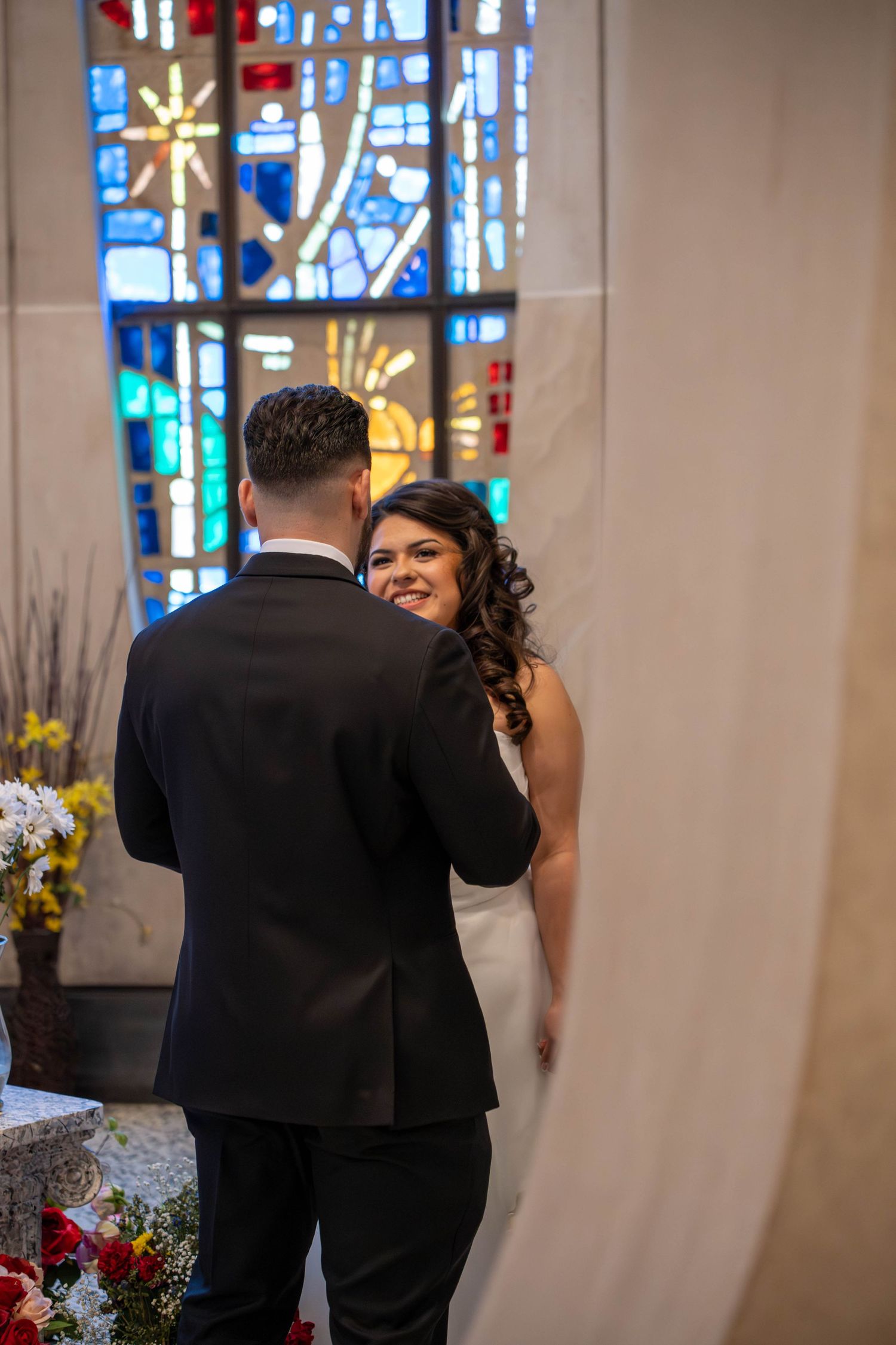 Bride smiling at groom during wedding ceremony in front of colorful stained glass church window.
