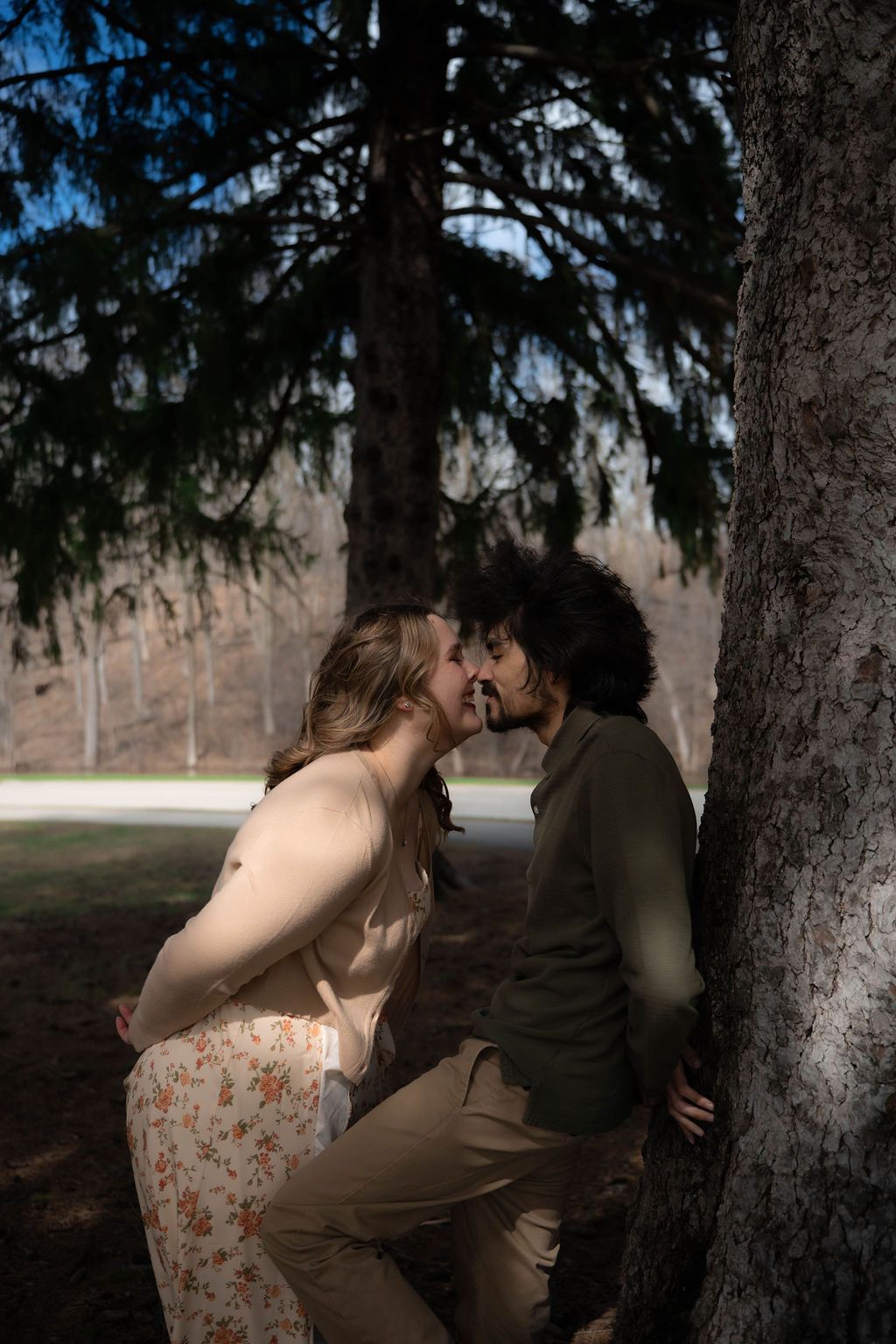 A couple sharing a romantic kiss beside a tree trunk in a sunlit park setting.
