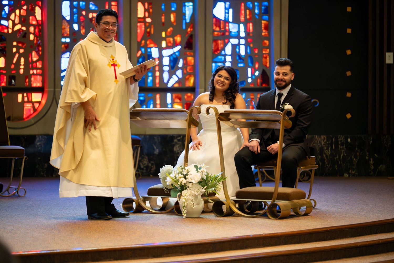 Priest officiating a wedding ceremony with bride and groom at church altar with colorful stained glass windows.
