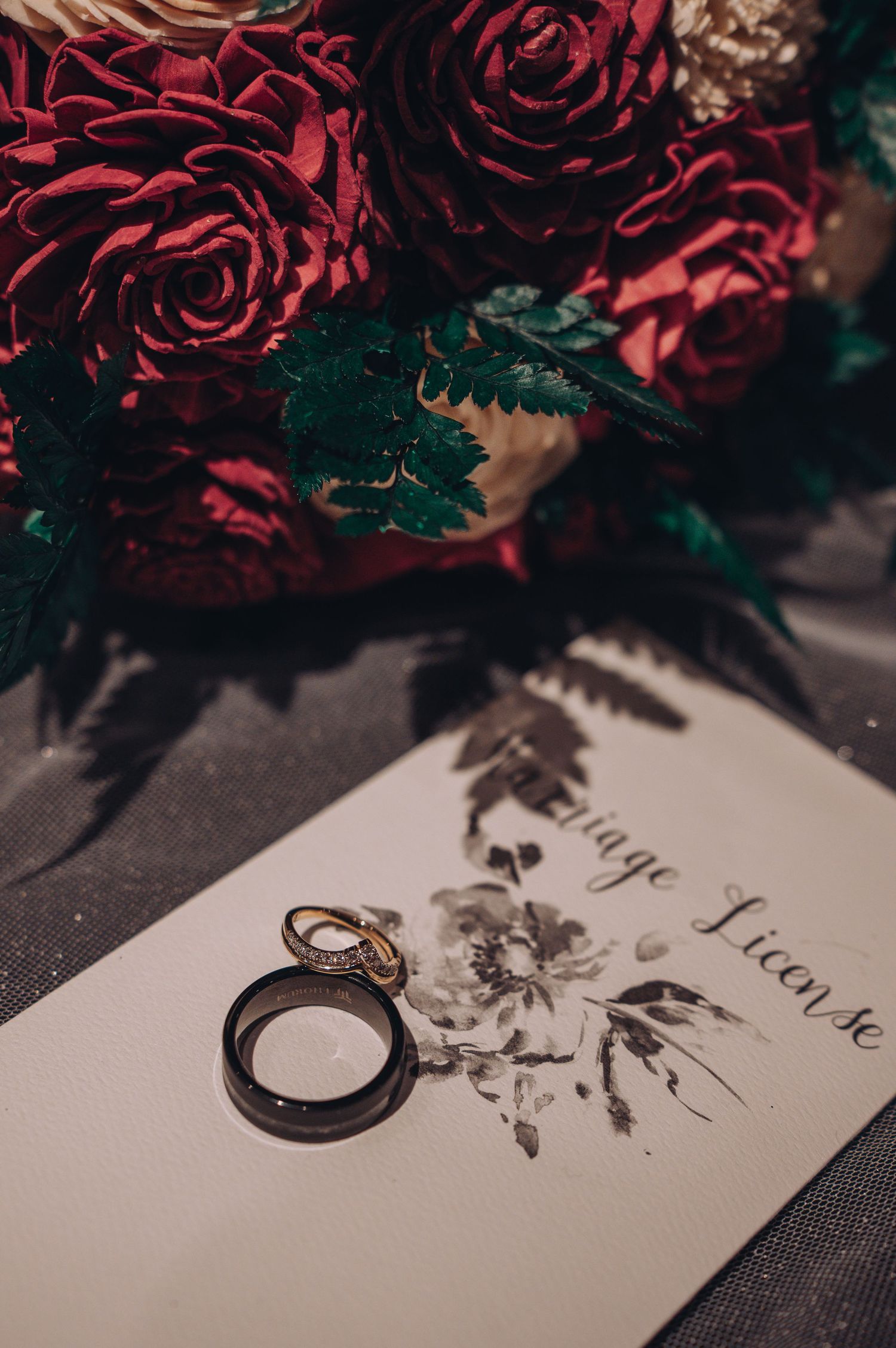 Wedding rings resting on marriage license beside red rose bouquet with greenery.