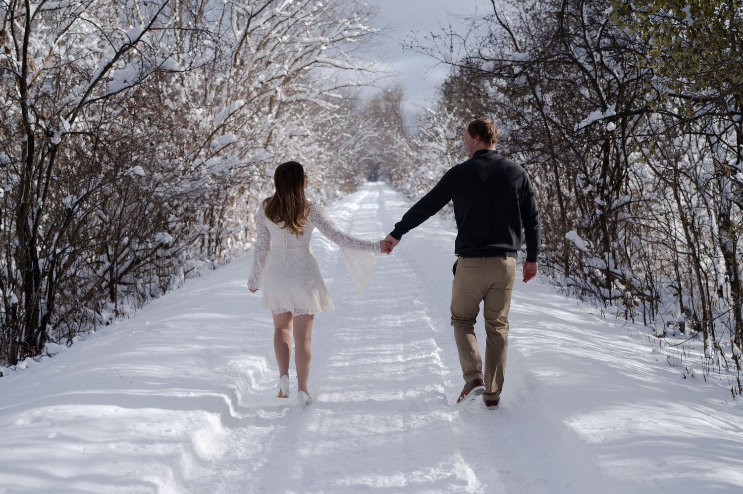 Couple holding hands walking down a snow-covered path lined with frost-covered trees in winter.