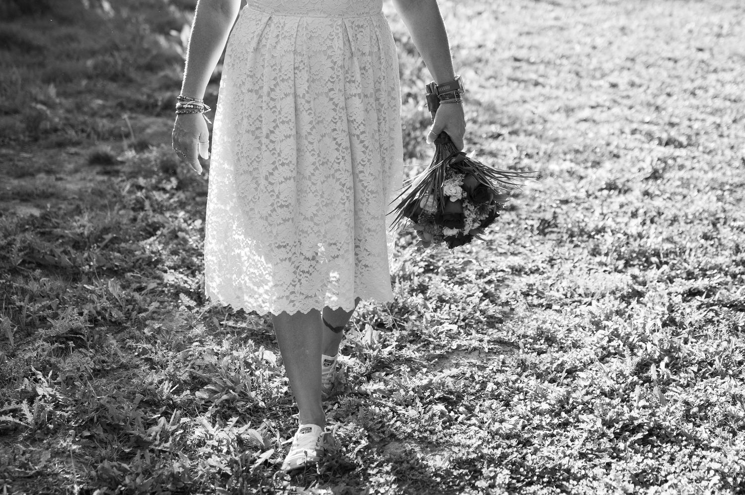 Black and white photo of bride in lace skirt holding bouquet in grassy field.