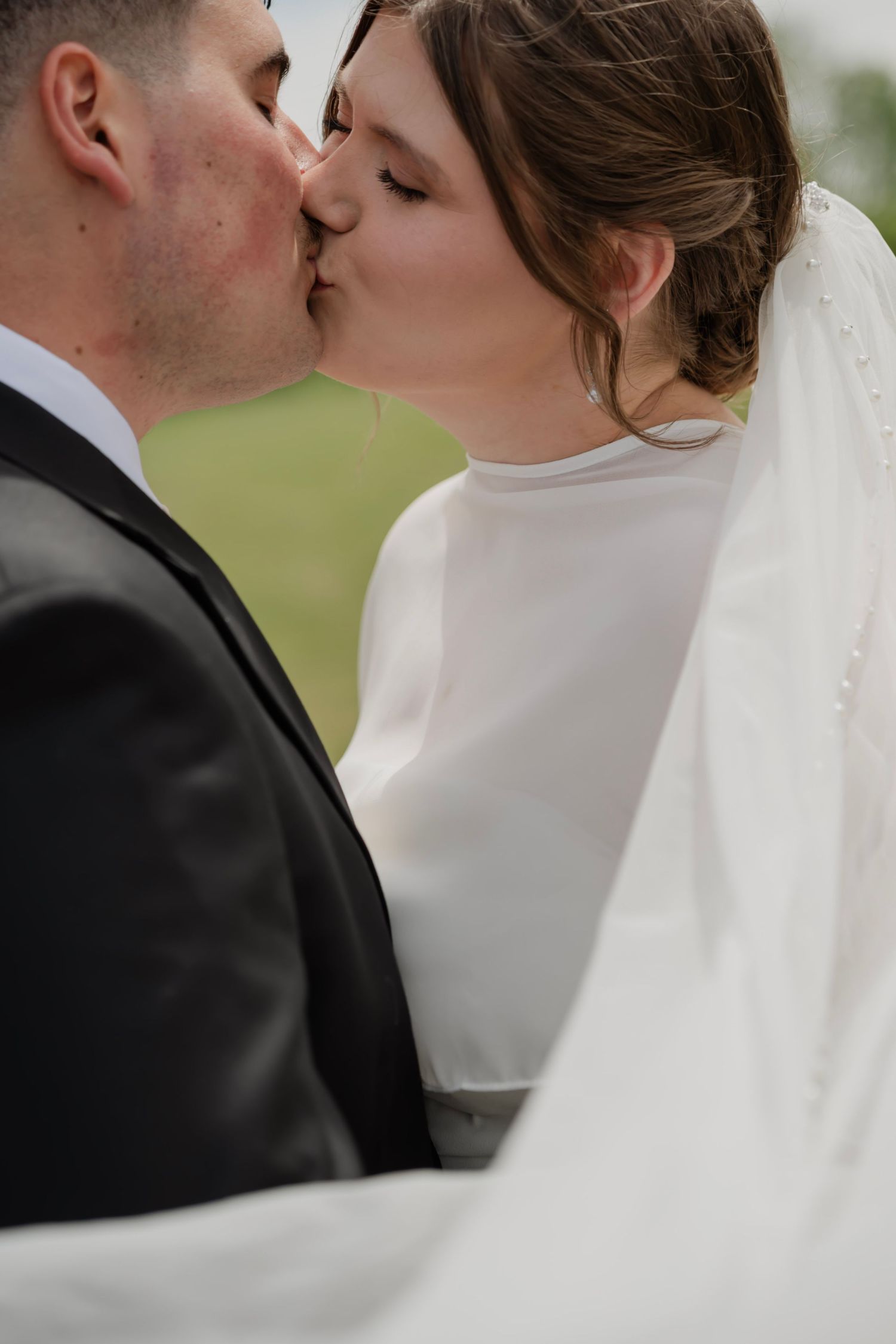A bride and groom share a romantic kiss outdoors, with her white veil flowing gracefully in the breeze.