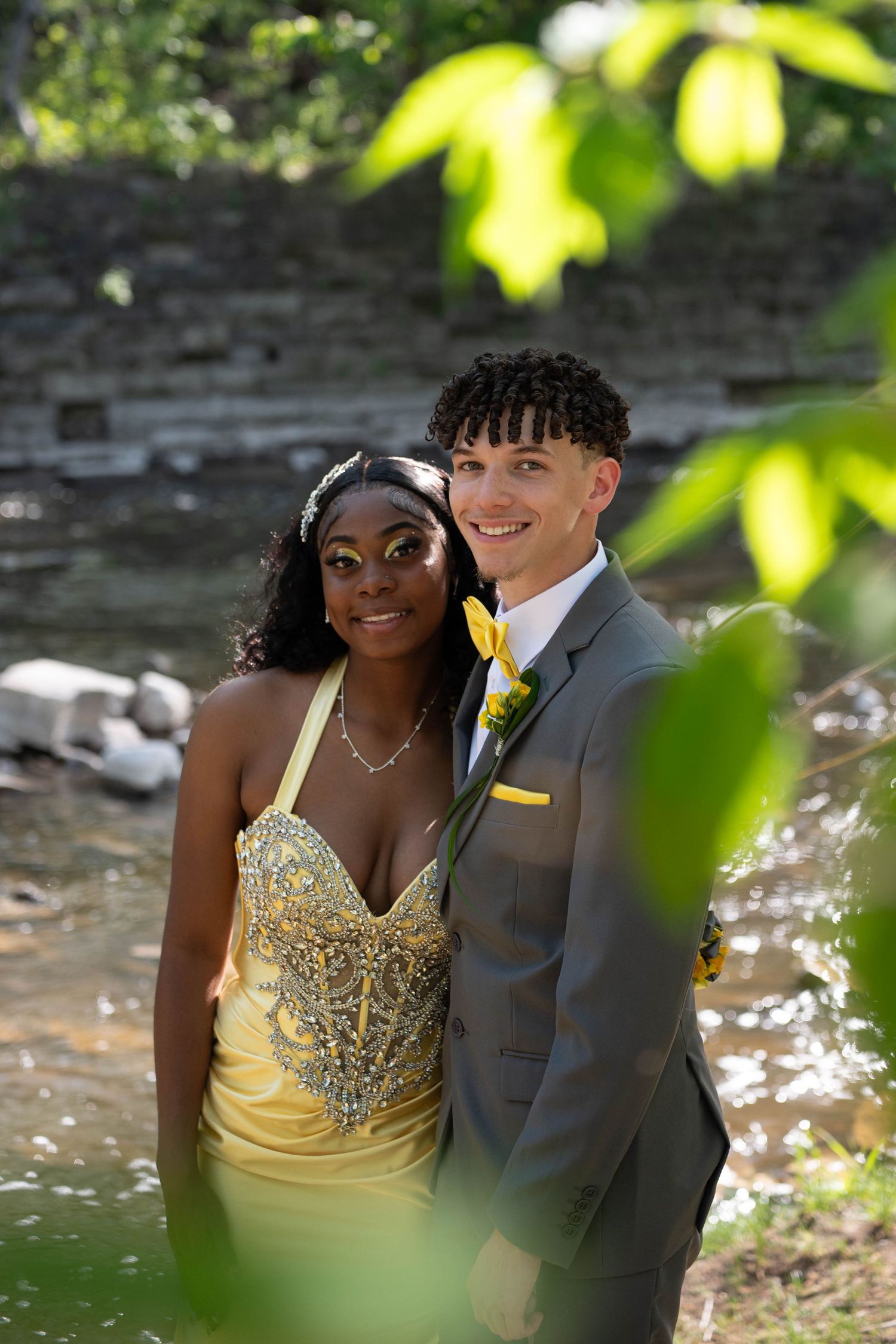 Prom couple in matching yellow outfits pose outdoors near a creek surrounded by lush green foliage.