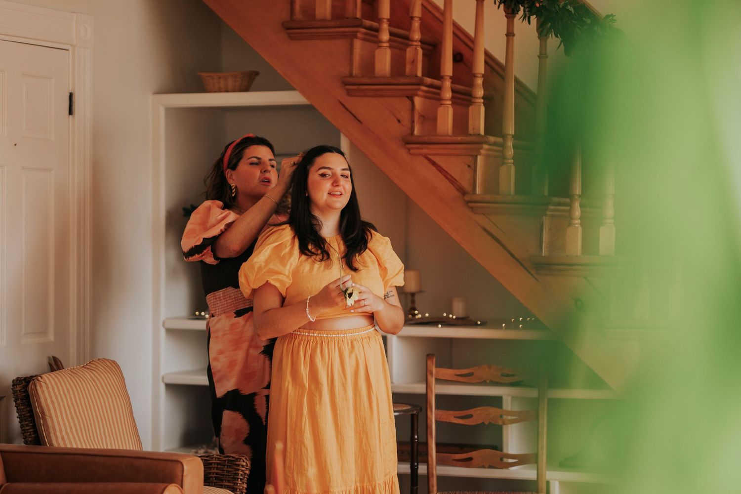 Two people stand in an indoor space with a wooden staircase, warm lighting and a yellow dress visible in frame.