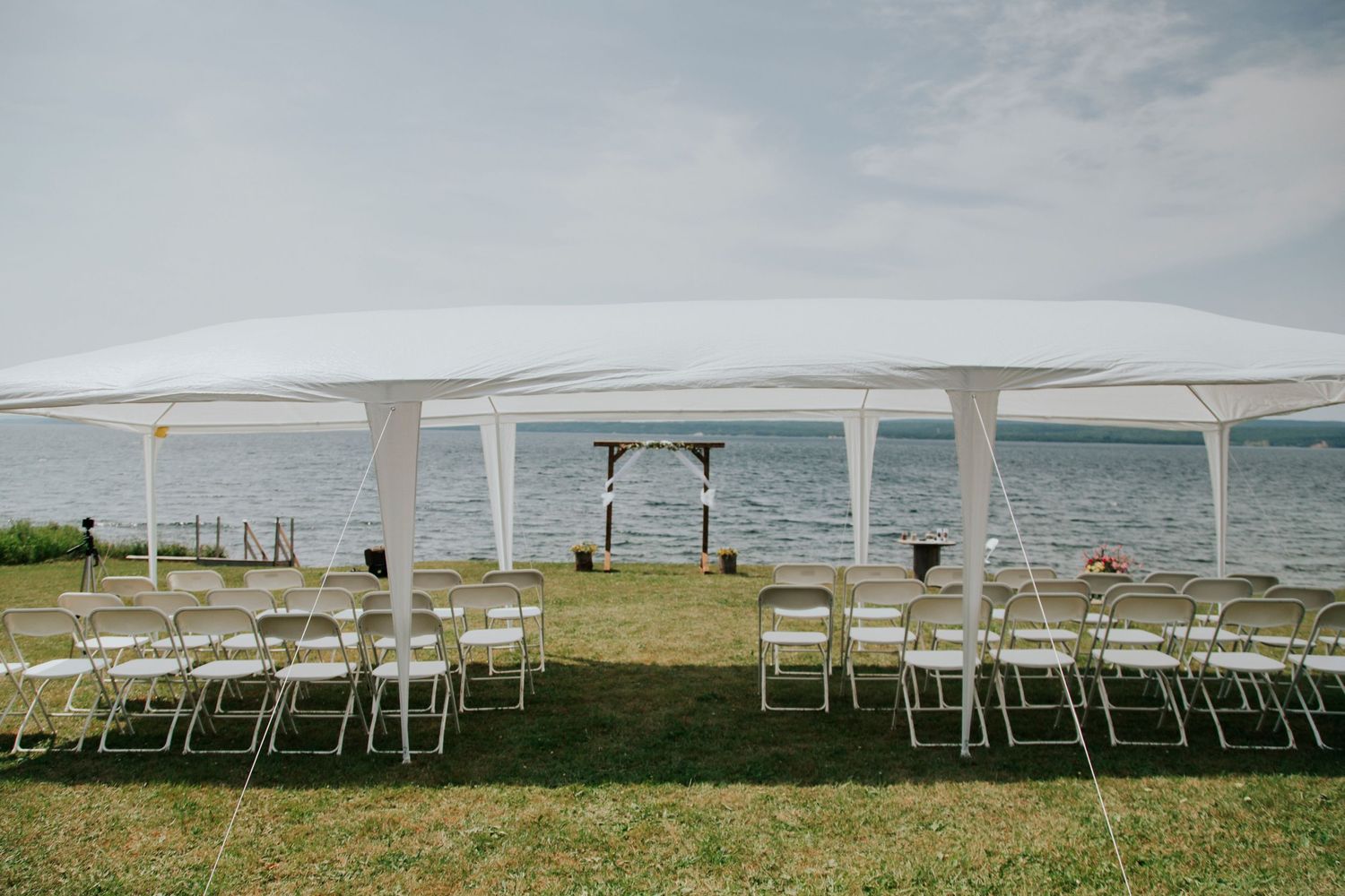 A white wedding tent set up on a grassy lawn overlooking the ocean with rows of folding chairs and an altar arch.