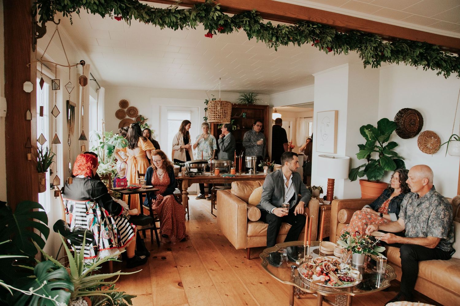 Guests mingling at a bohemian indoor wedding reception with lush greenery decor and warm wooden floors.