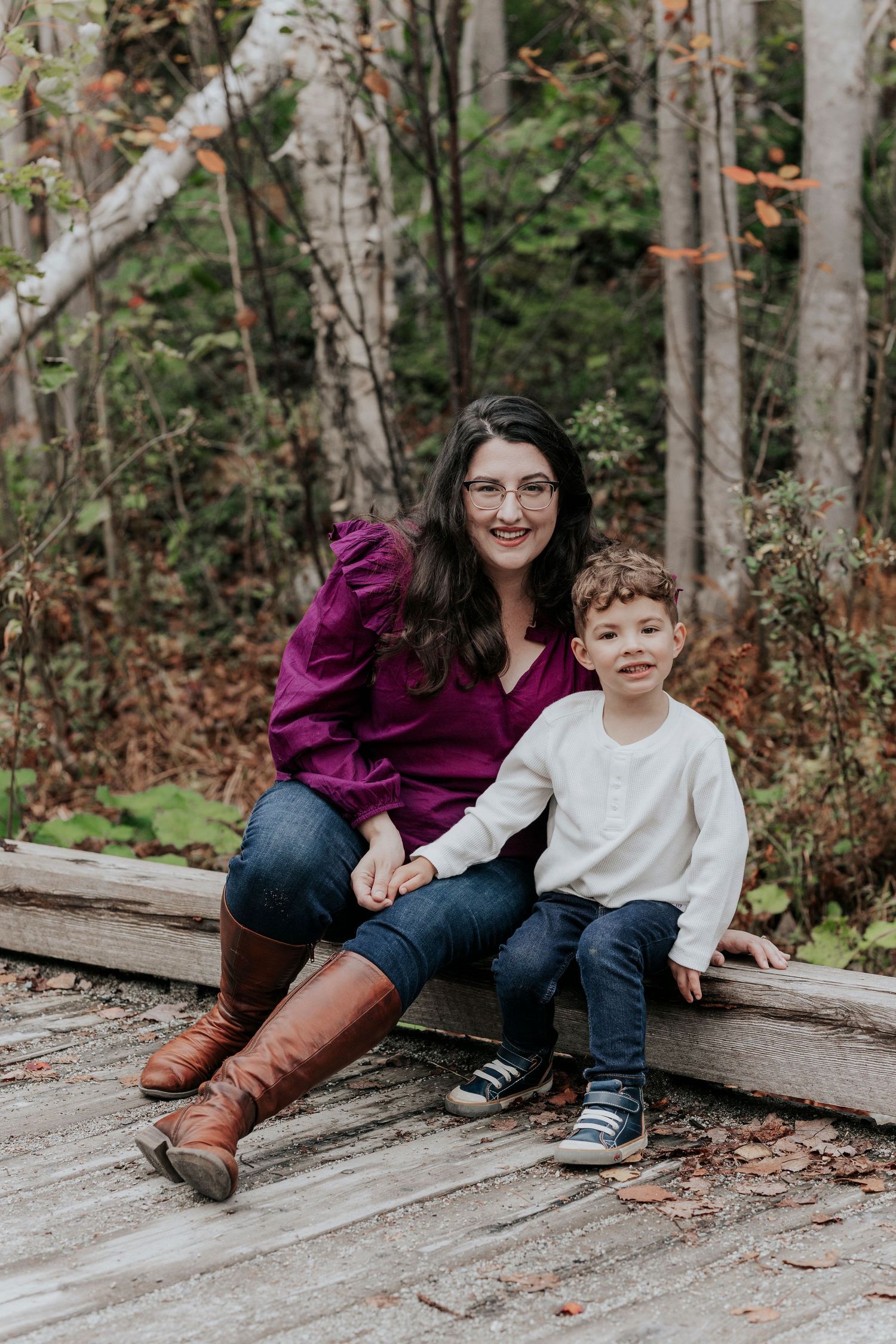 Smiling mother with glasses and young son sitting on wooden boards in autumn forest.