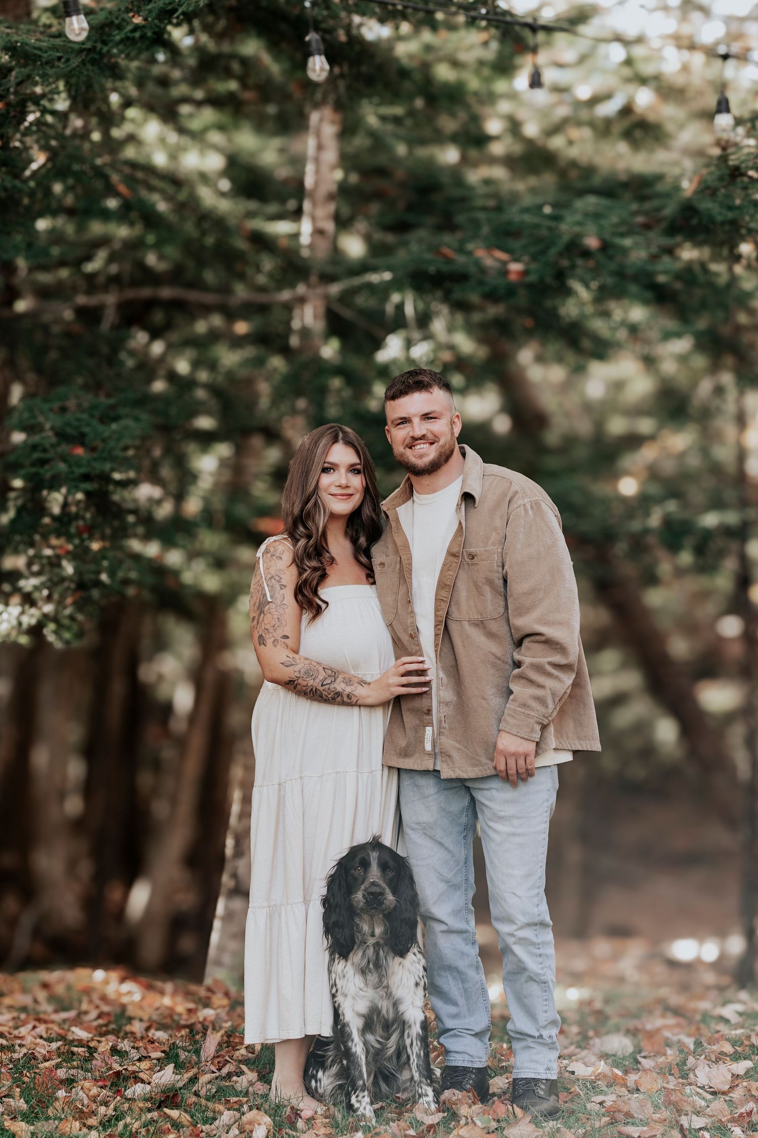 Couple poses with their spotted dog among autumn trees, woman in white dress, man in tan jacket and jeans.