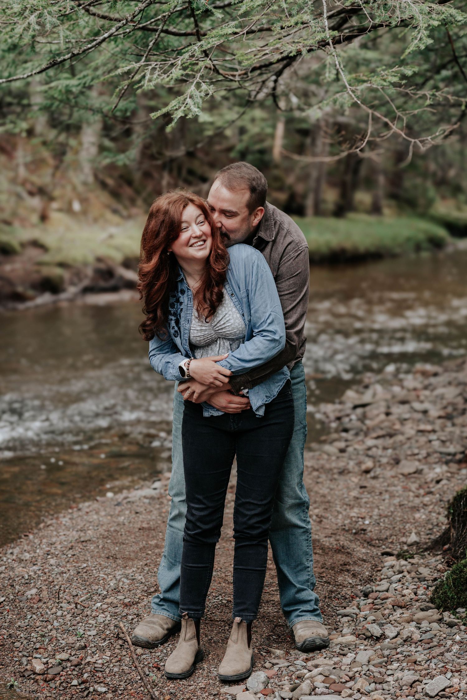 Couple embracing by a forest creek, man kissing woman's cheek as she smiles in denim jacket.