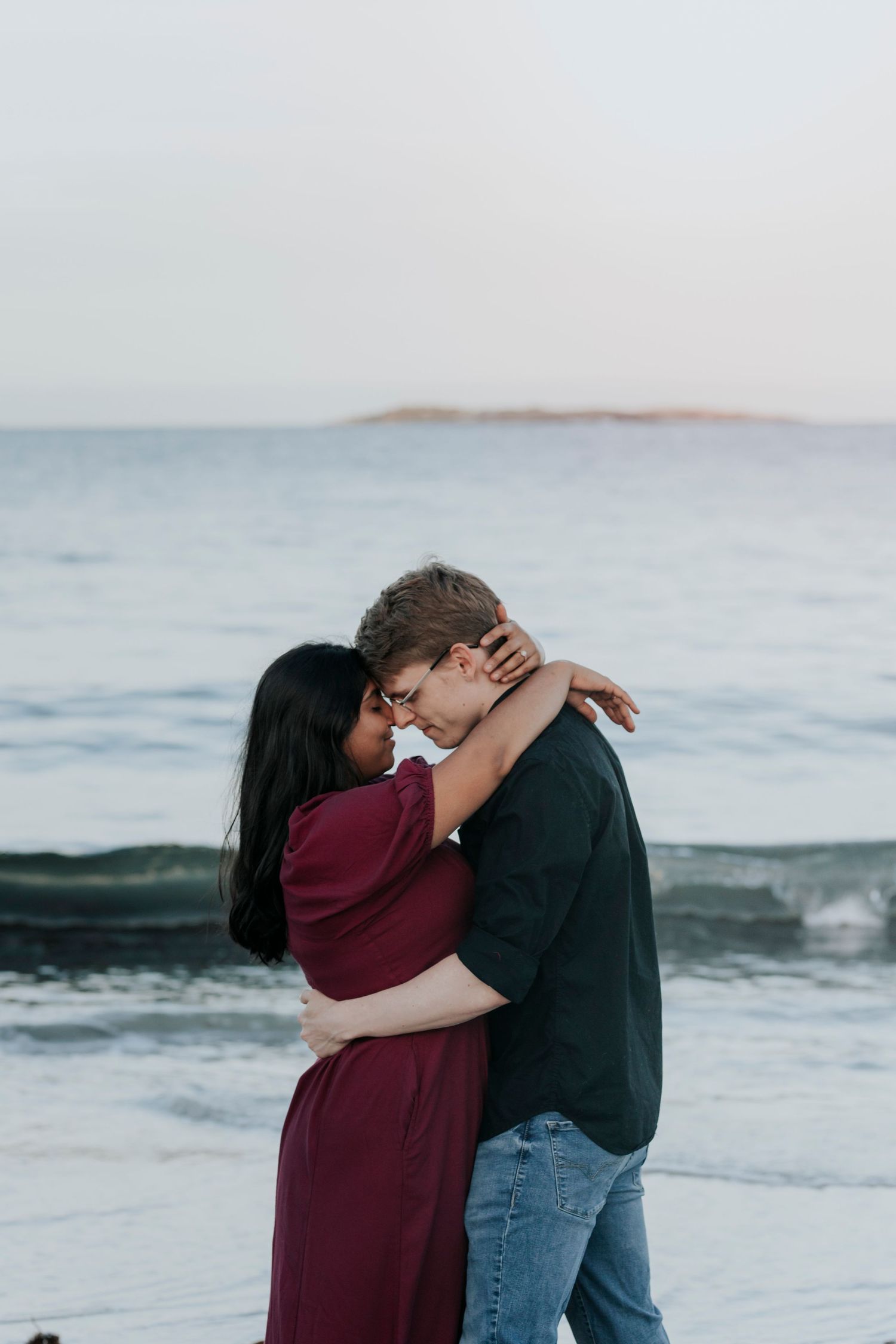 Couple shares an intimate embrace on a beach at dusk with ocean waves in the background.