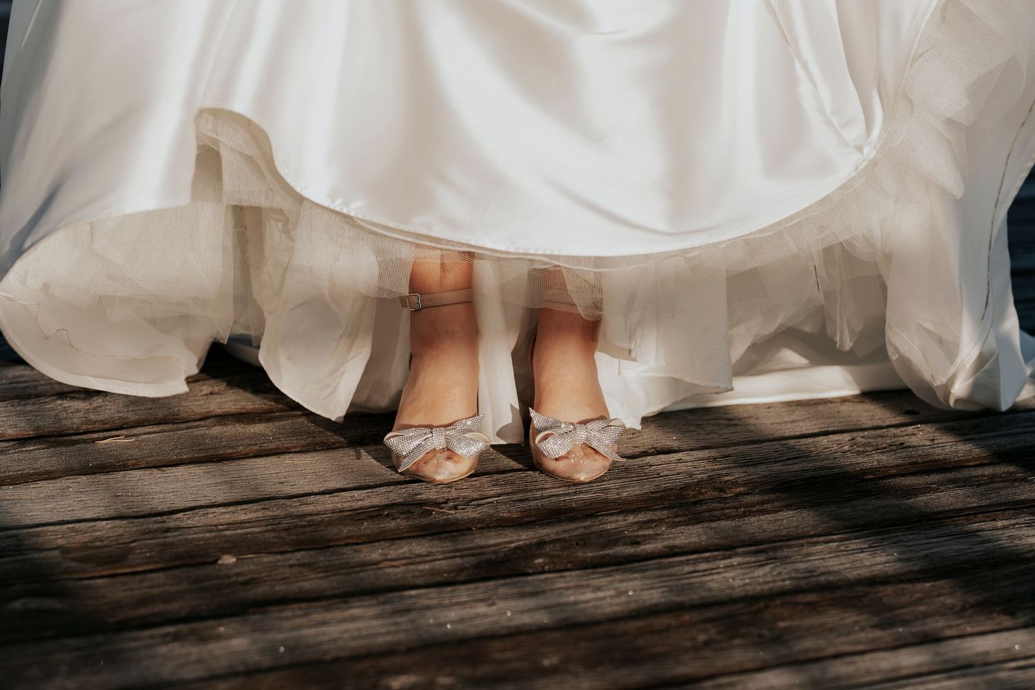 Close-up view of bridal shoes peeking out from under a flowing white wedding dress on wooden deck planks.