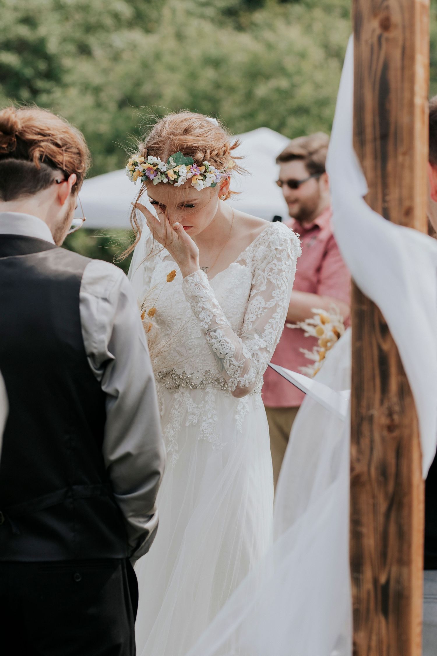 A bride in a lace wedding dress and floral crown shares an emotional moment during an outdoor ceremony.