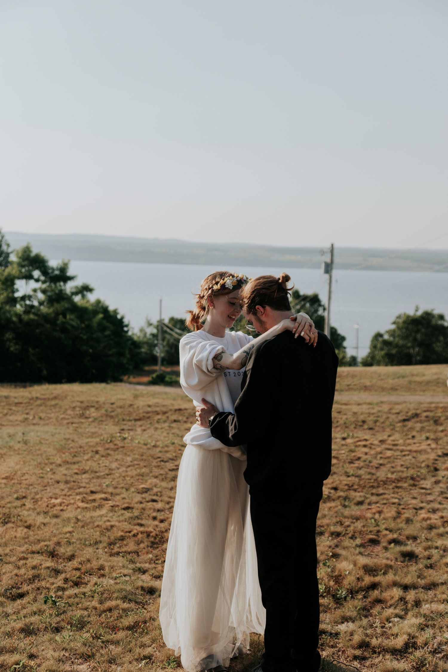 A couple embraces on a hilltop overlooking Bras d'Or lake, with the bride wearing a flowing white dress and floral crown.
