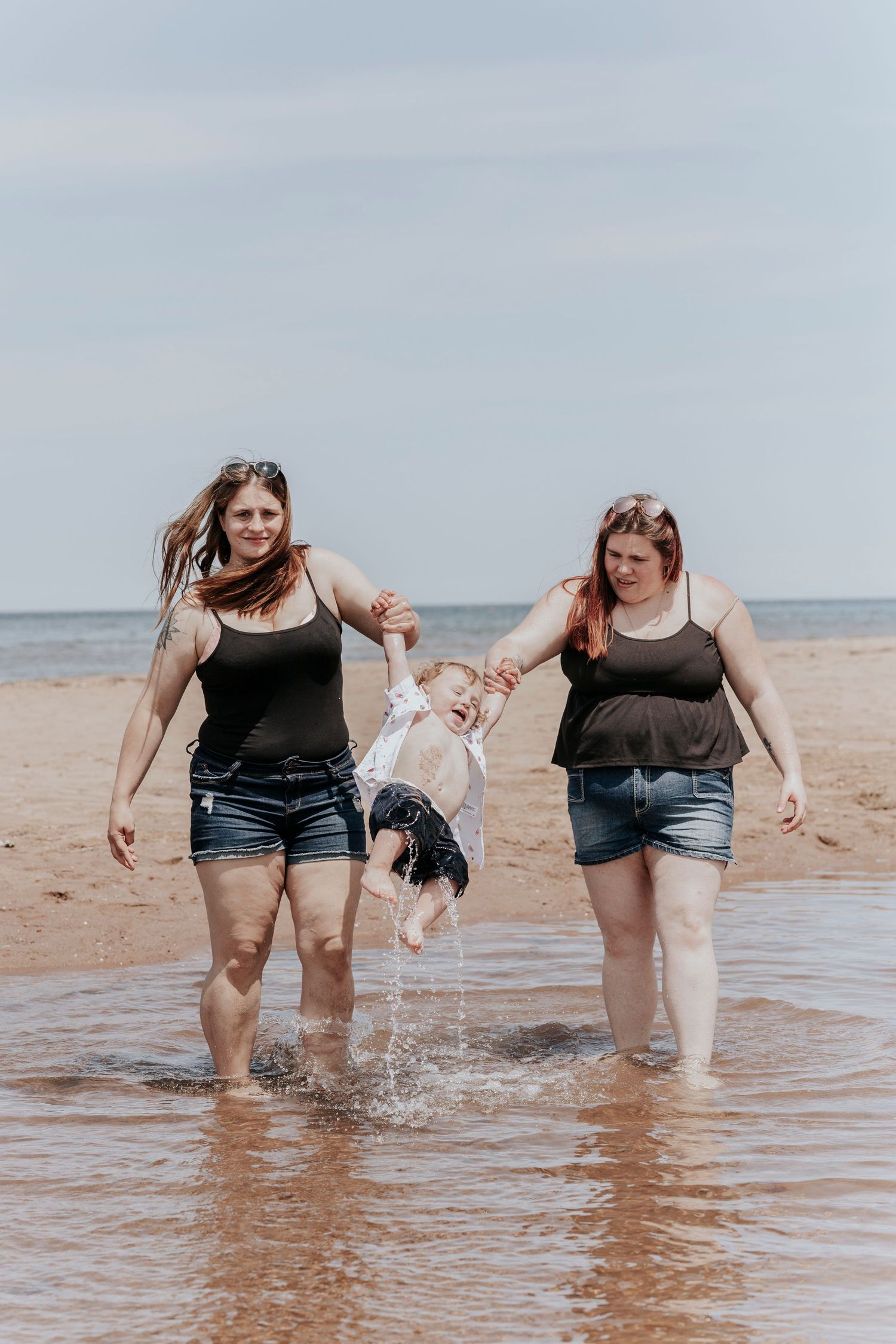 Two women swinging a laughing toddler over ocean waves on a sandy beach.