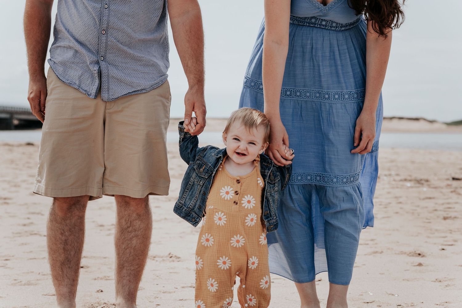 Toddler girl in yellow daisy romper and denim jacket holding parents hands on a sandy beach.
