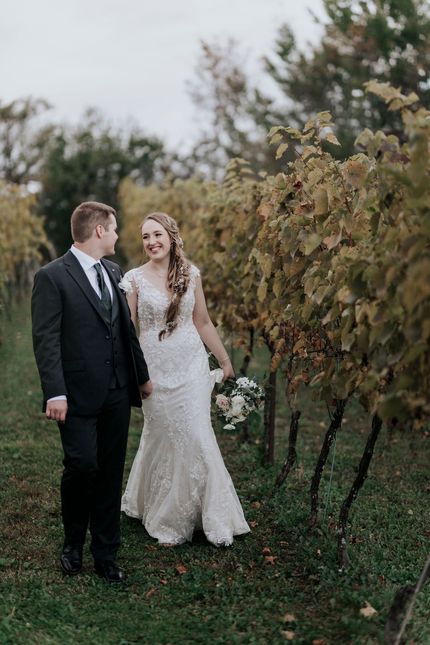 Smiling bride in lace gown holds bouquet while walking with groom through autumn vineyard rows.
