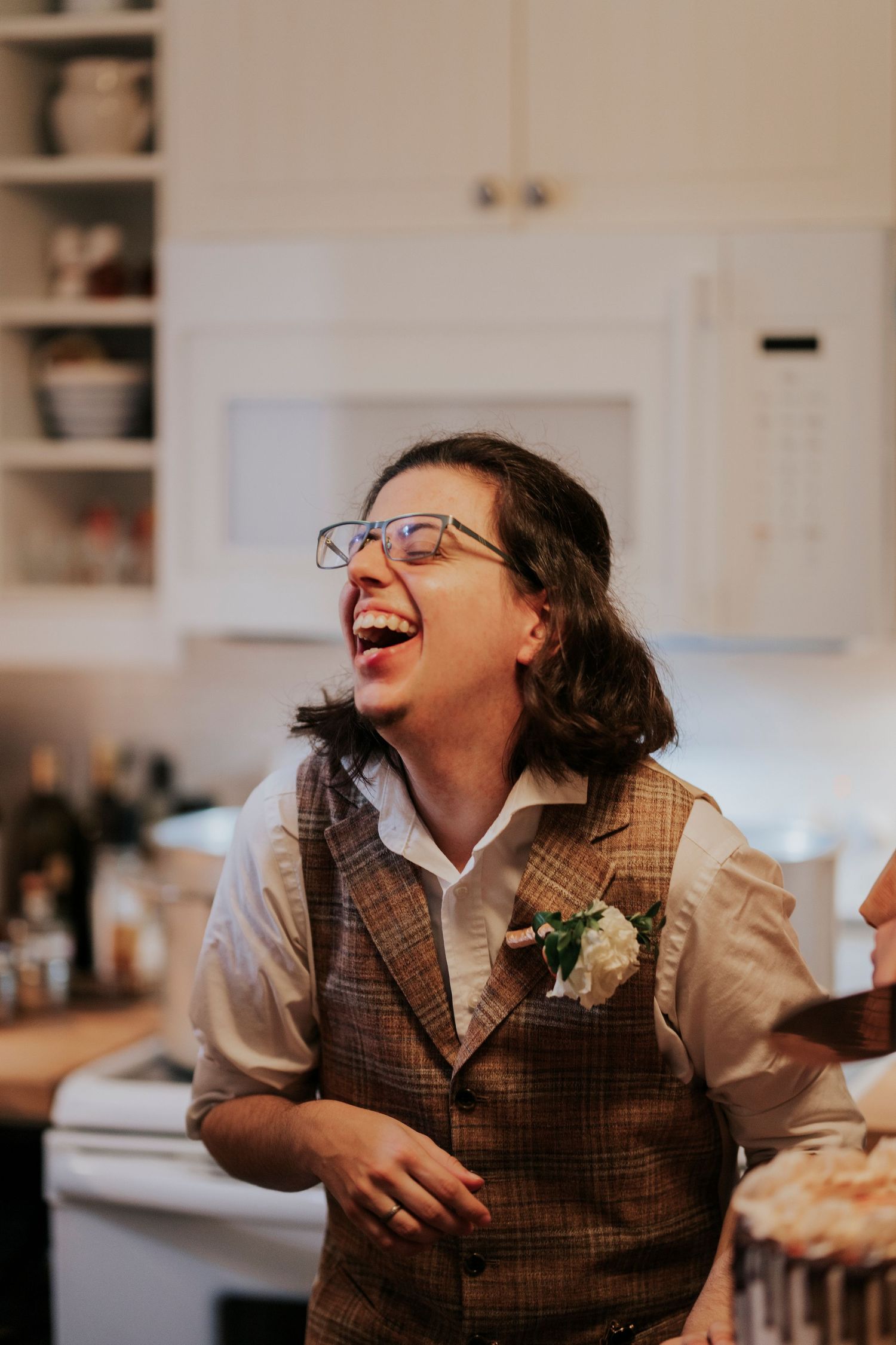 A laughing person wearing glasses and a tweed vest with a boutonniere in a wedding kitchen setting.