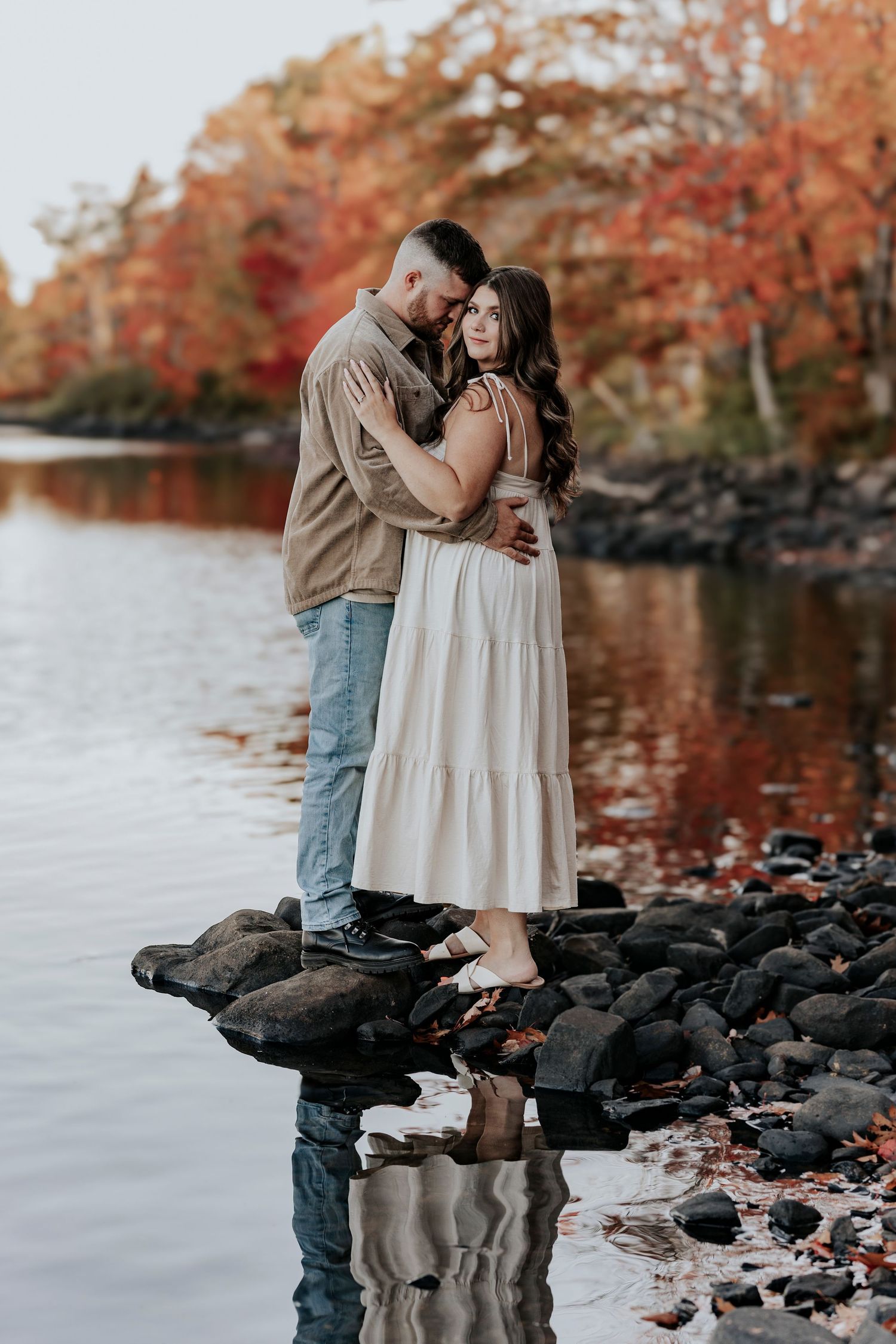 Couple stands on rocks by a lake surrounded by vibrant fall foliage, reflecting in the calm water.