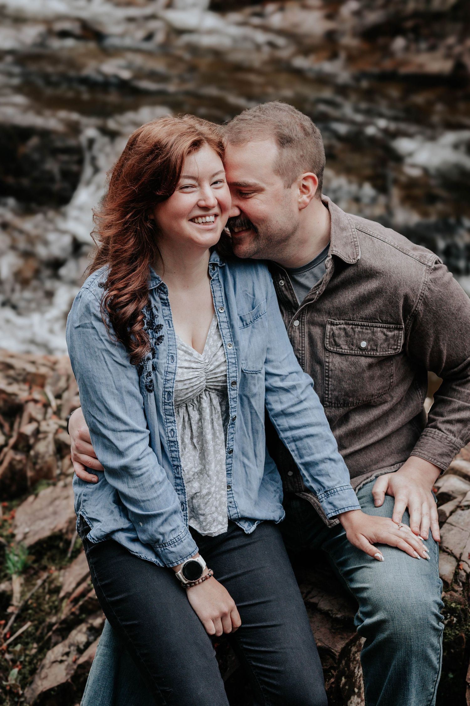 Smiling redhead woman and man sitting on rocks by waterfall, sharing a close romantic moment.