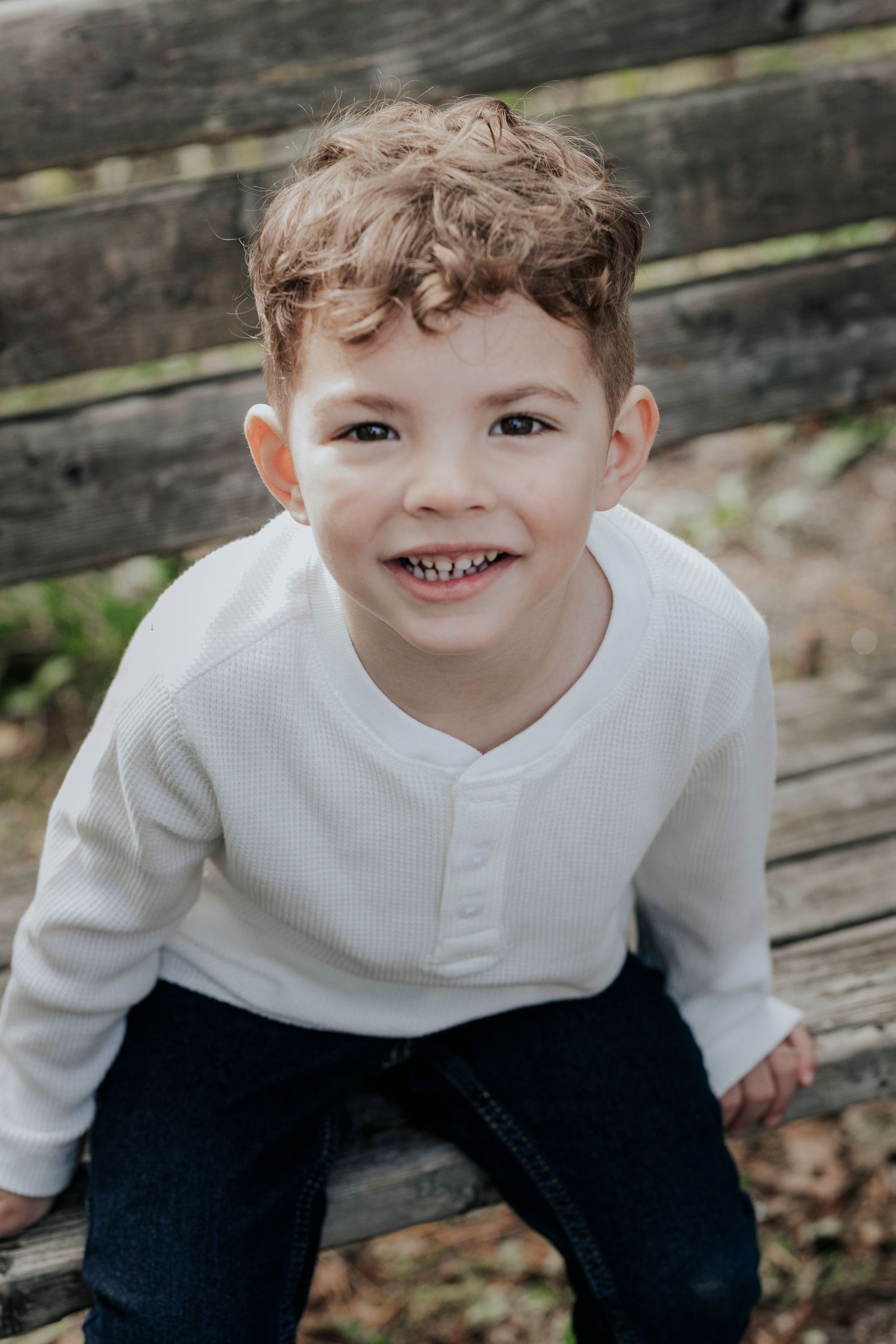 Cute curly-haired young boy in white shirt smiling while sitting on wooden boards outdoors.