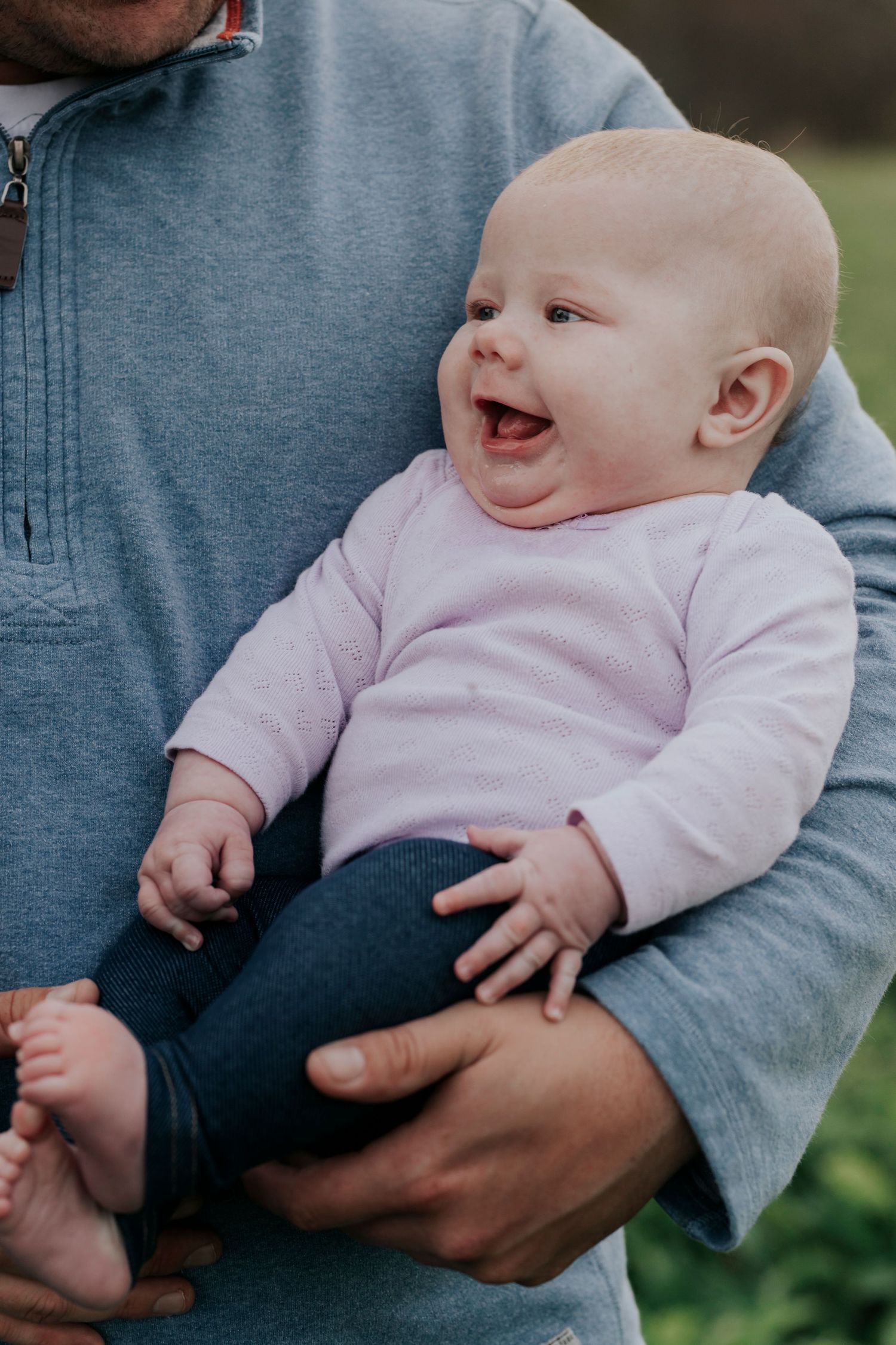 Laughing baby in pink outfit being held by father outdoors.