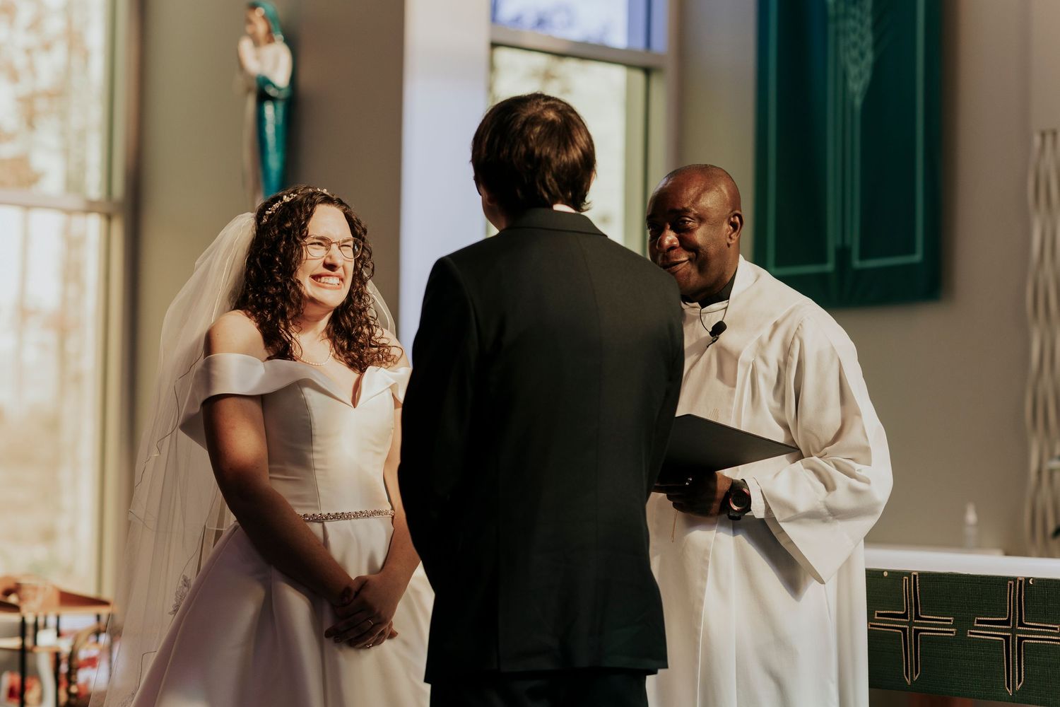 A wedding ceremony takes place in a bright church interior as a couple stands before an officiant at the altar.