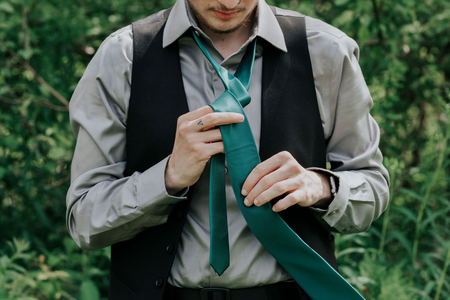 The groom wearing a dress shirt and vest adjusts their green tie against a leafy outdoor background.