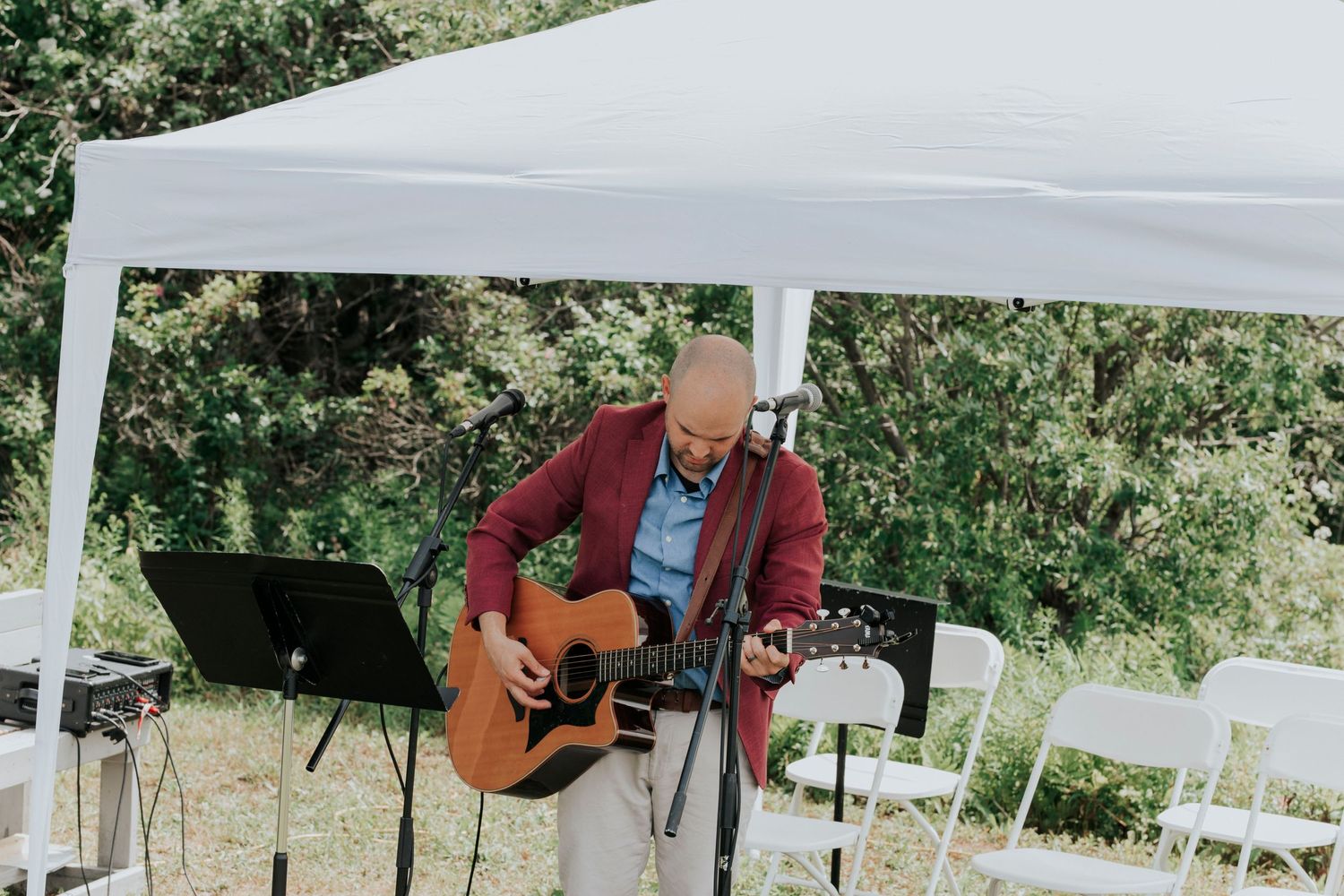 Musician performing with acoustic guitar under white canopy tent outdoors in natural setting.