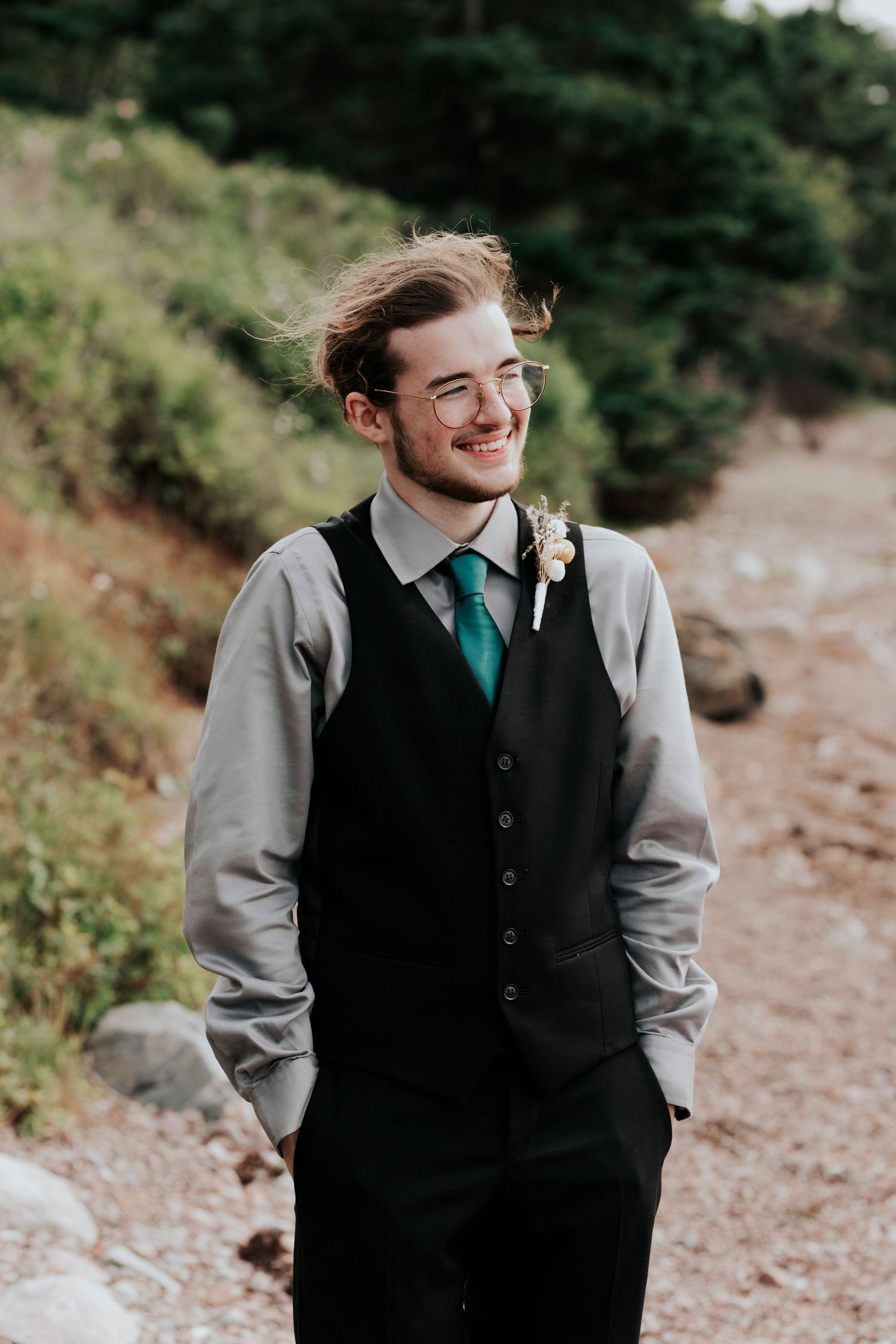 A groom in formal attire with a black vest and emerald green tie smiles while standing on a beach in Cape Breton