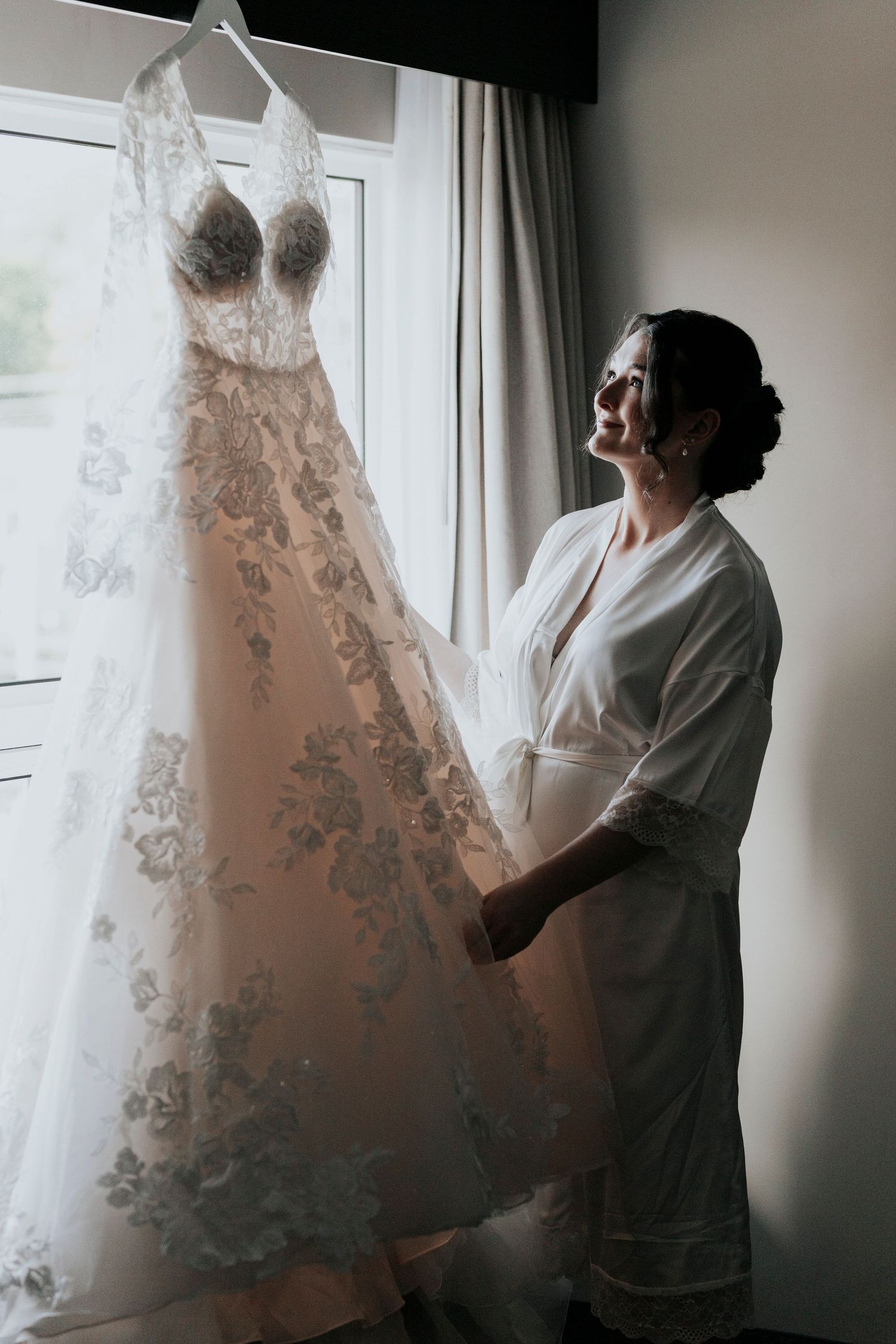 A wedding dress with intricate lace detailing hangs by a window as someone in white robe admires it.