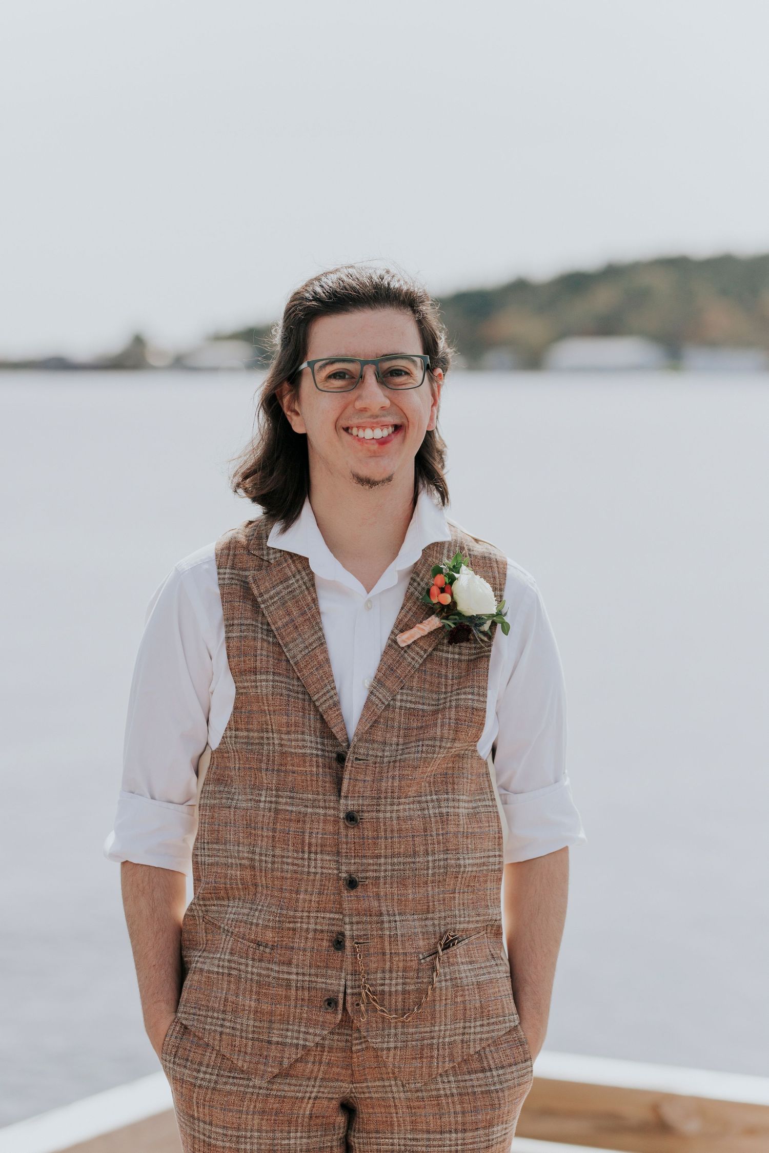 The groom wearing glasses and a plaid tweed vest with a boutonniere poses outdoors against a waterfront background.