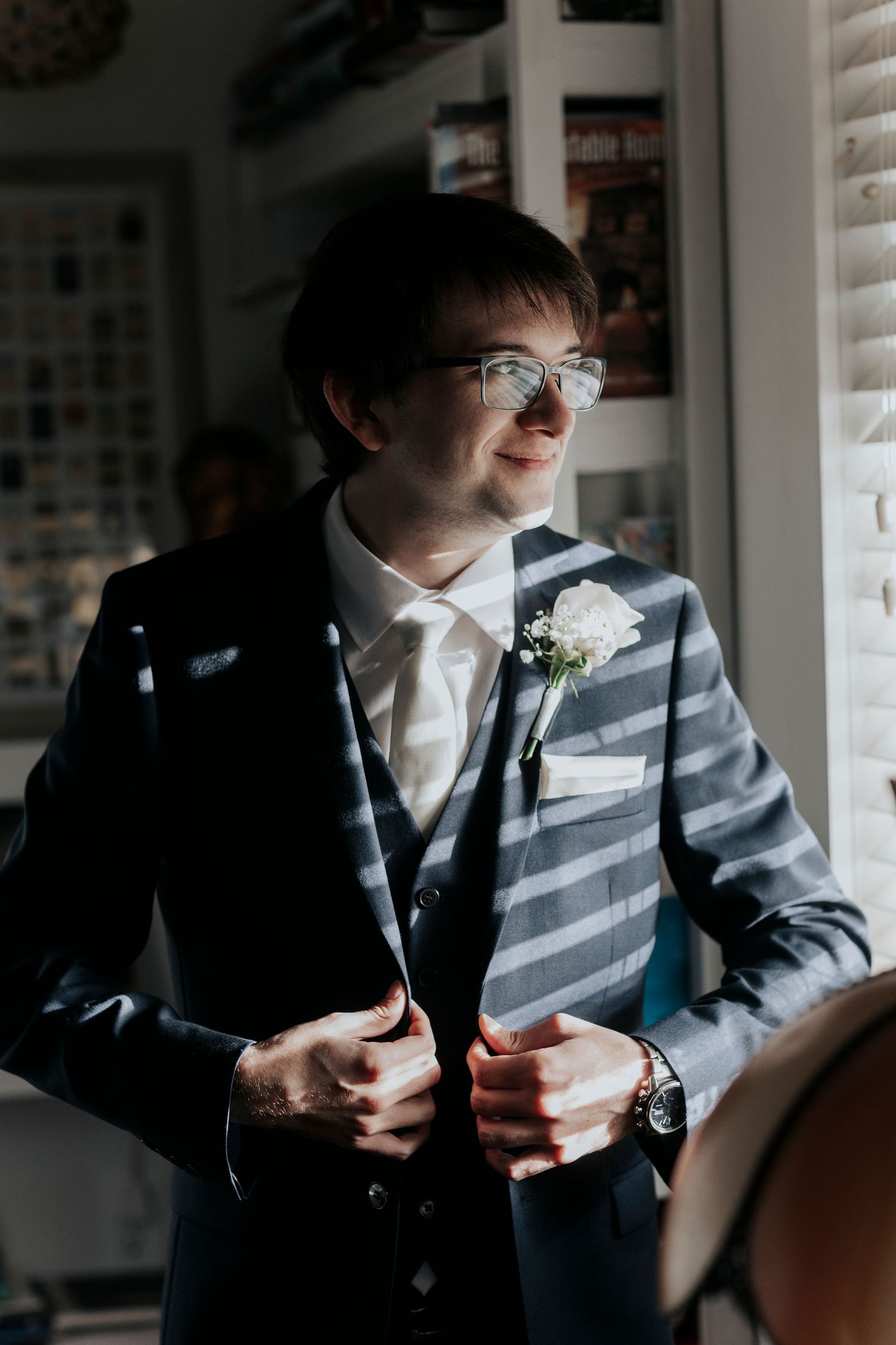 A groom in a navy suit and white boutonniere stands near a window with sunlight casting striped shadows through blinds.