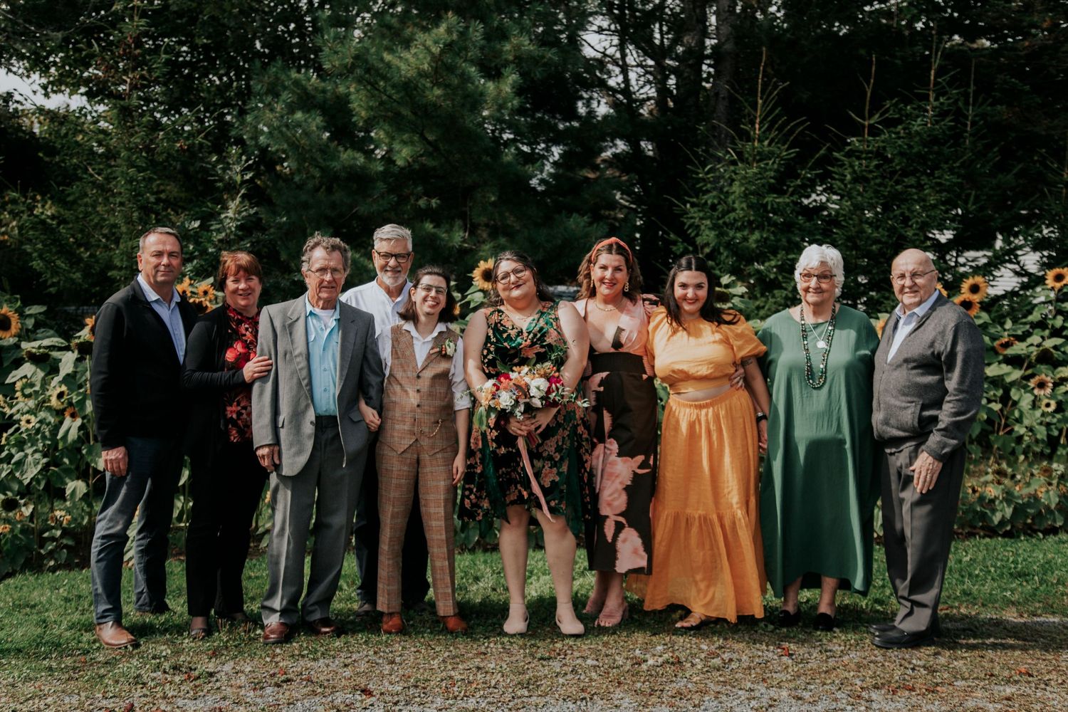 A group of dressed-up wedding guests gather outdoors for a photo in a lush garden setting.