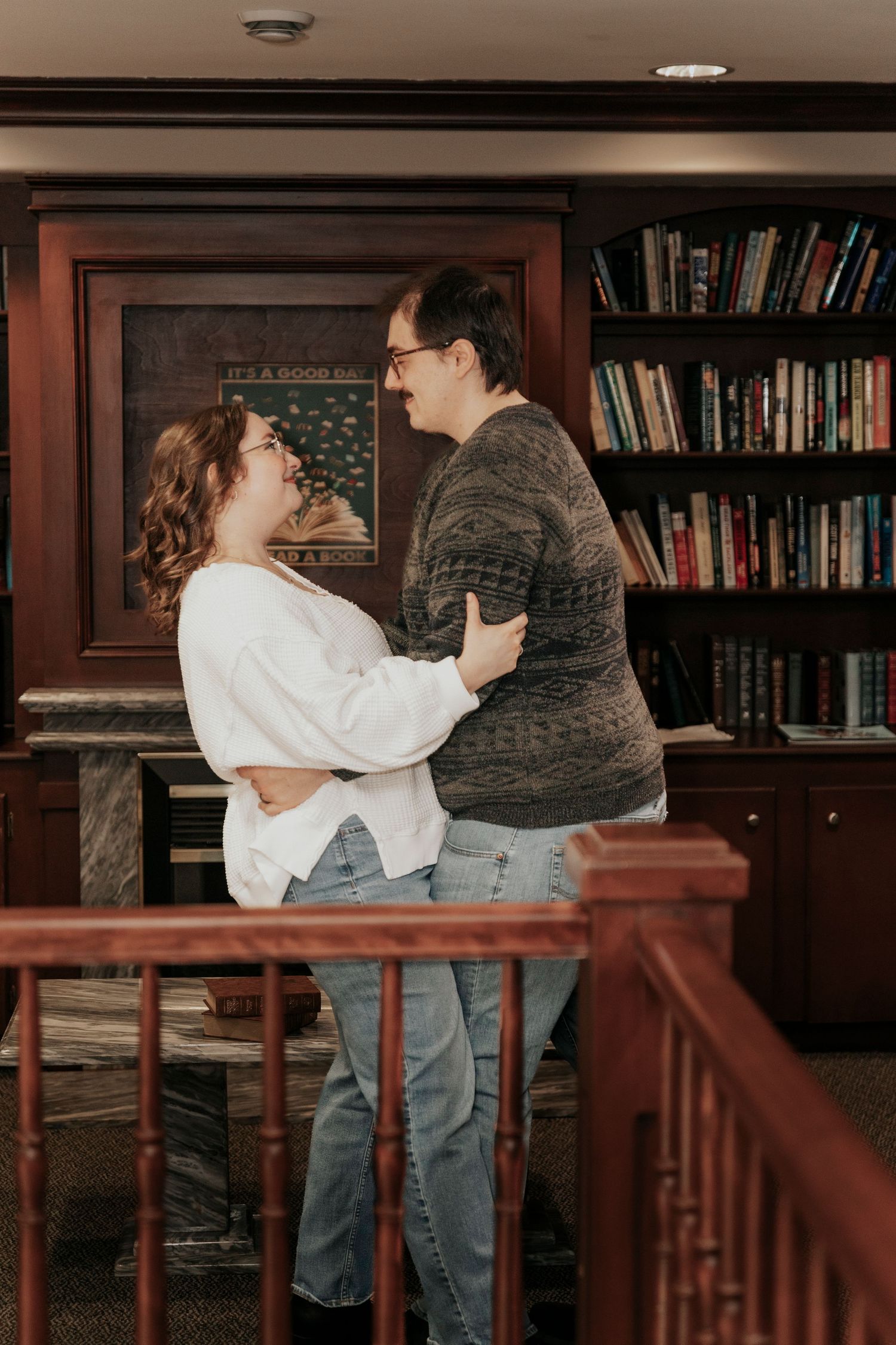 Couple embracing and gazing at each other in a cozy library with wooden bookshelves.
