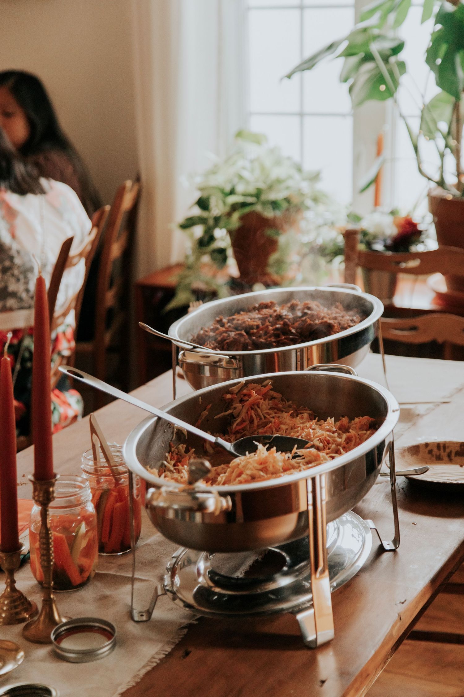 Buffet setup with chafing dishes of food and red candles on wooden table with houseplants in background.