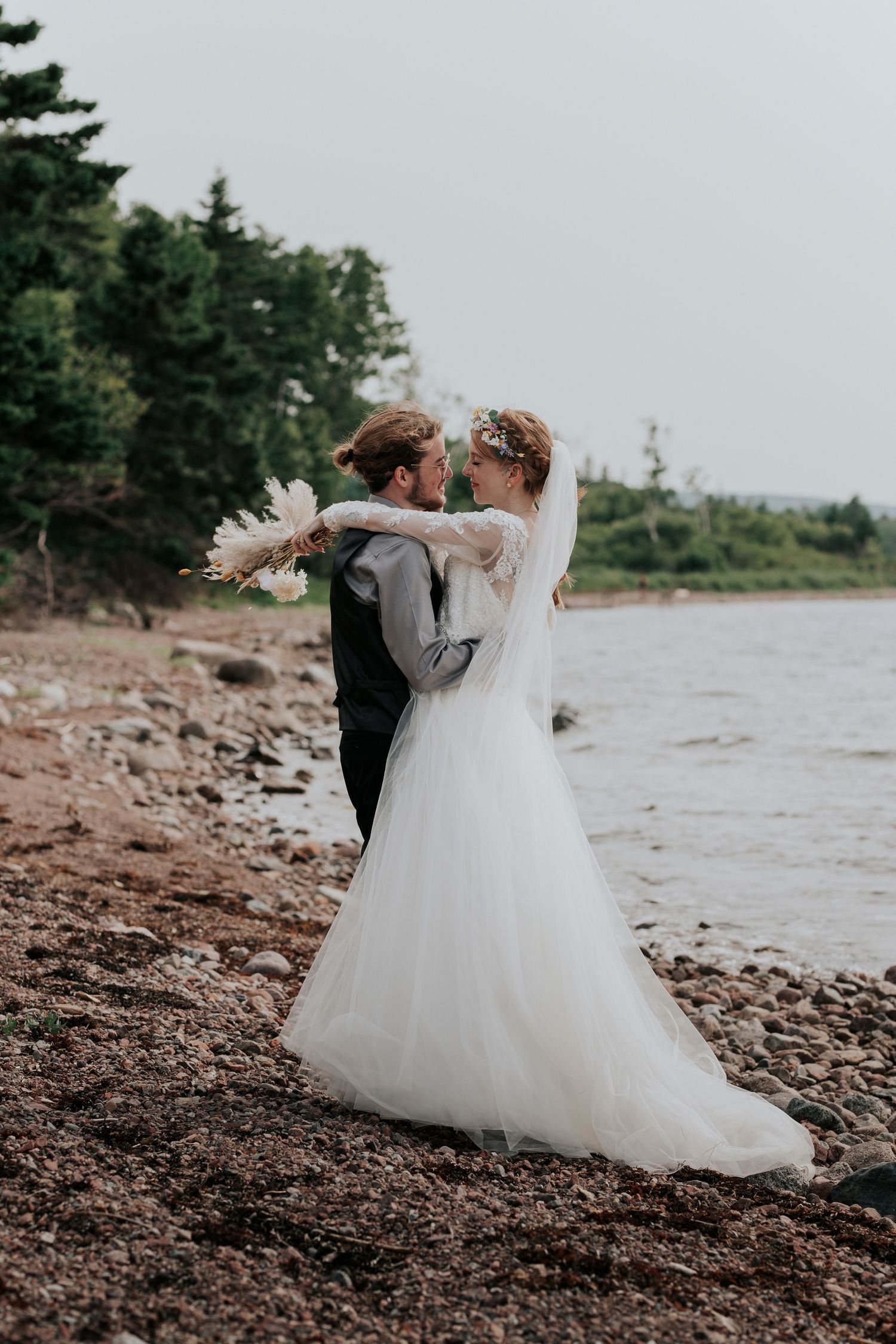 Bride and groom embrace on a rocky shoreline, bride in flowing white gown holding pampas grass bouquet.