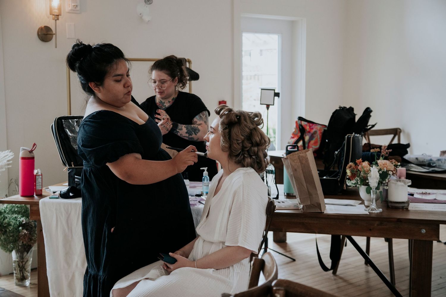 Professional makeup artist and hairstylist work on getting their client ready in a bright interior setting.