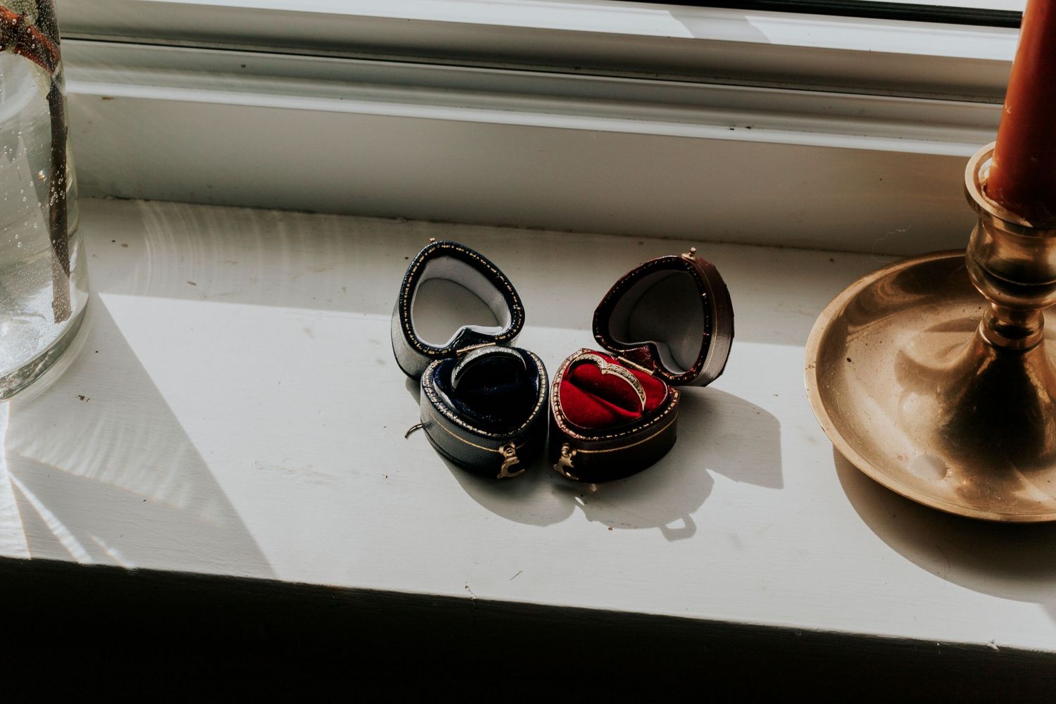 Black and red ring boxes resting on a white windowsill beside a brass candlestick holder.