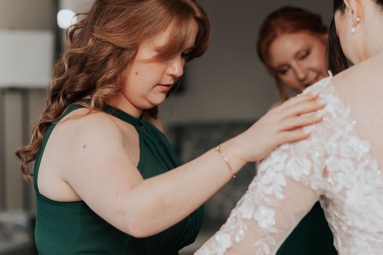 A bridesmaid in a dark green dress helps adjust the lace sleeve of a wedding gown.