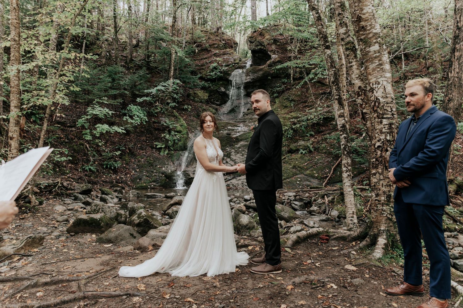 Bride and groom exchange vows in a forest elopement ceremony with a waterfall backdrop and witness present.