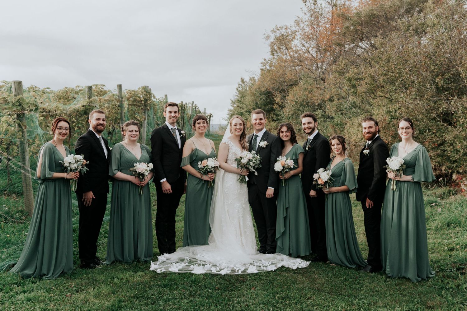 A wedding party in sage green dresses and black suits posing in an autumn vineyard.
