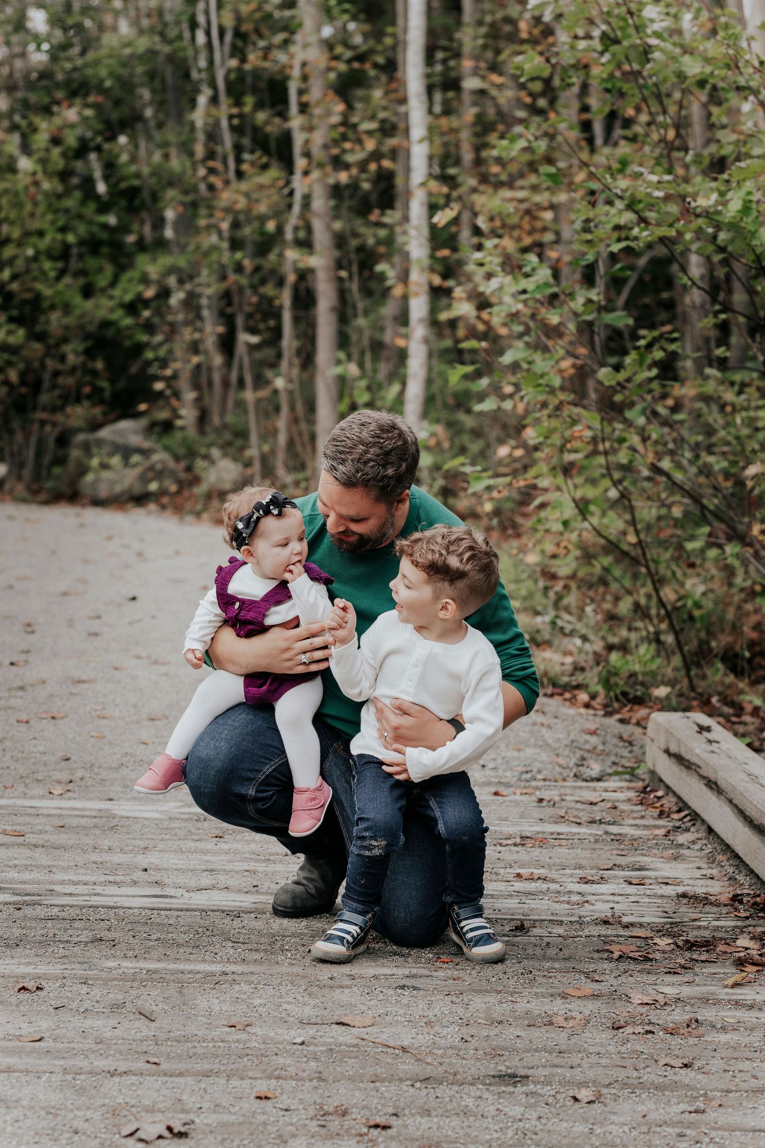 Father in green sweater crouching with baby girl and young son on woodland path.