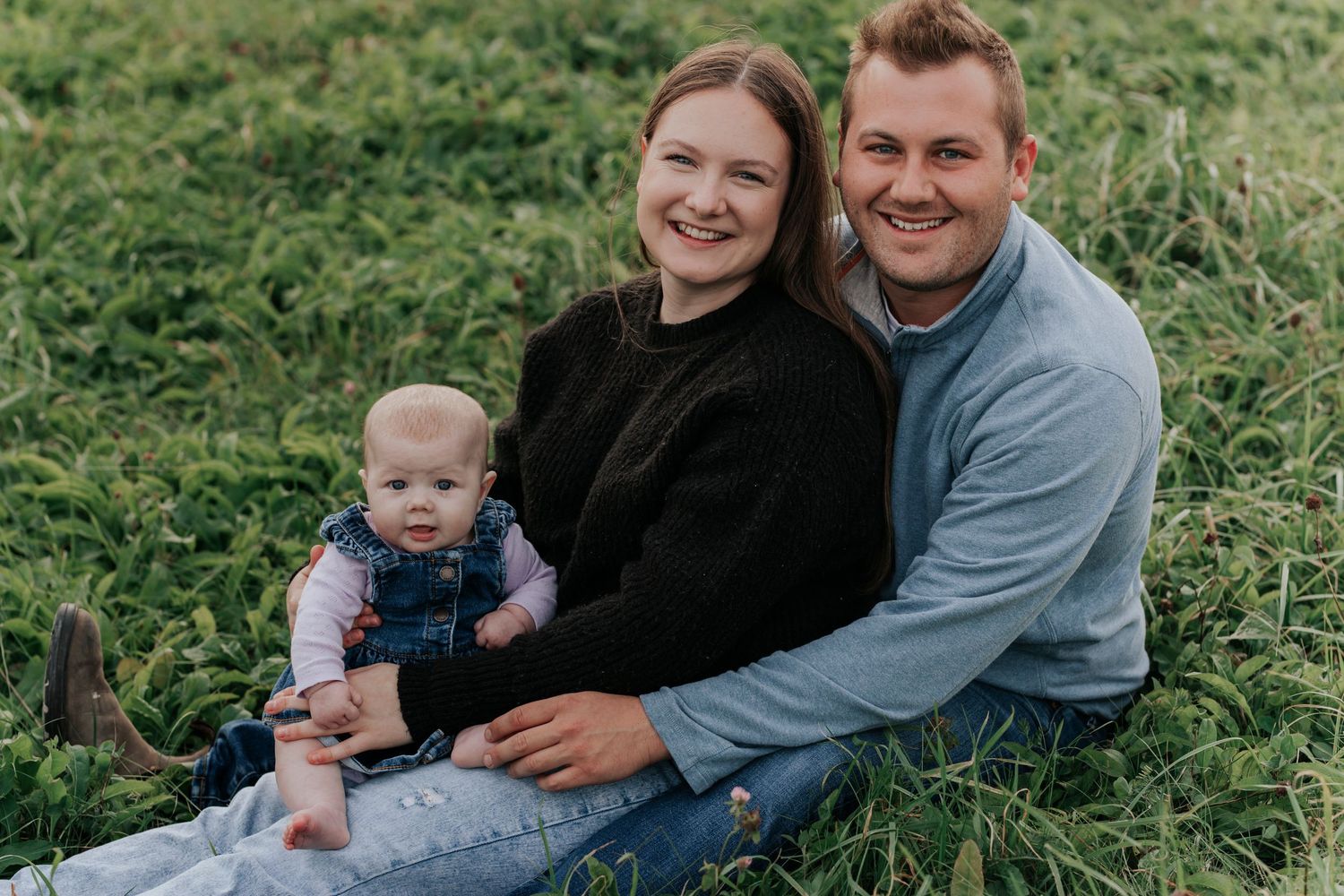 Smiling couple sitting in grass with baby in denim overalls.
