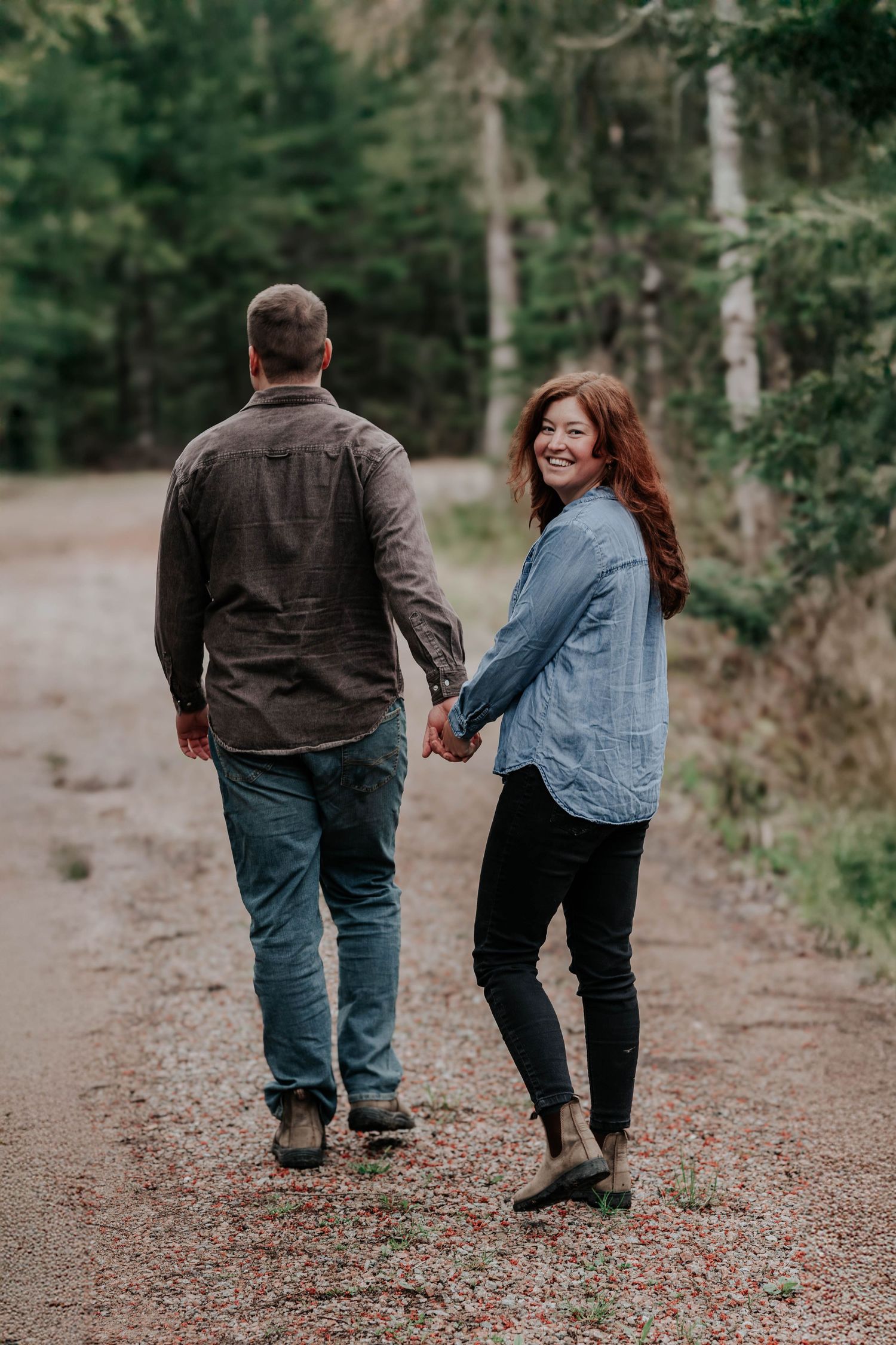 Couple walking hand in hand on forest path, woman looking back smiling over her shoulder.