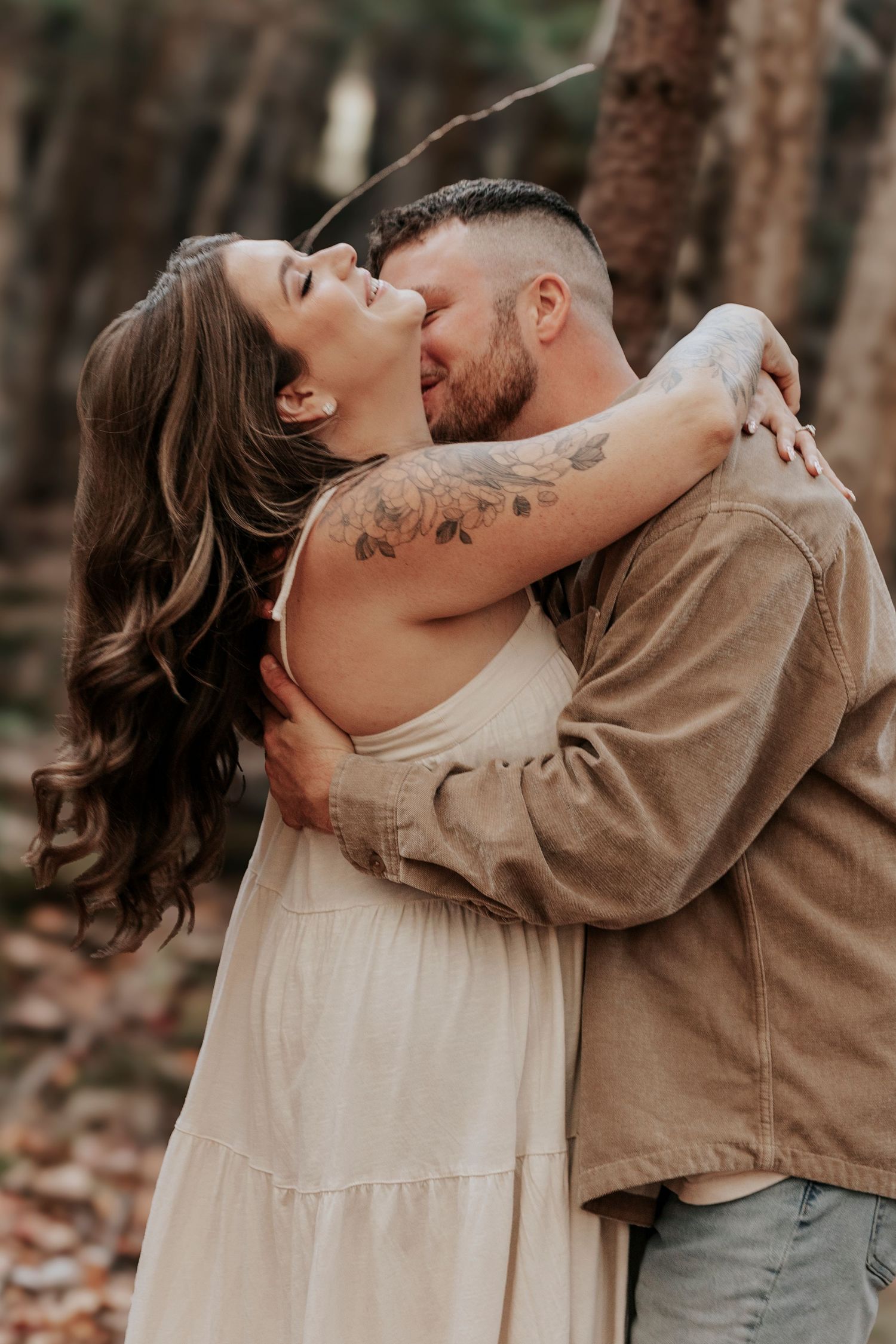 Couple shares a tender embrace in an autumn forest, woman with floral tattoos in white dress laughing joyfully.