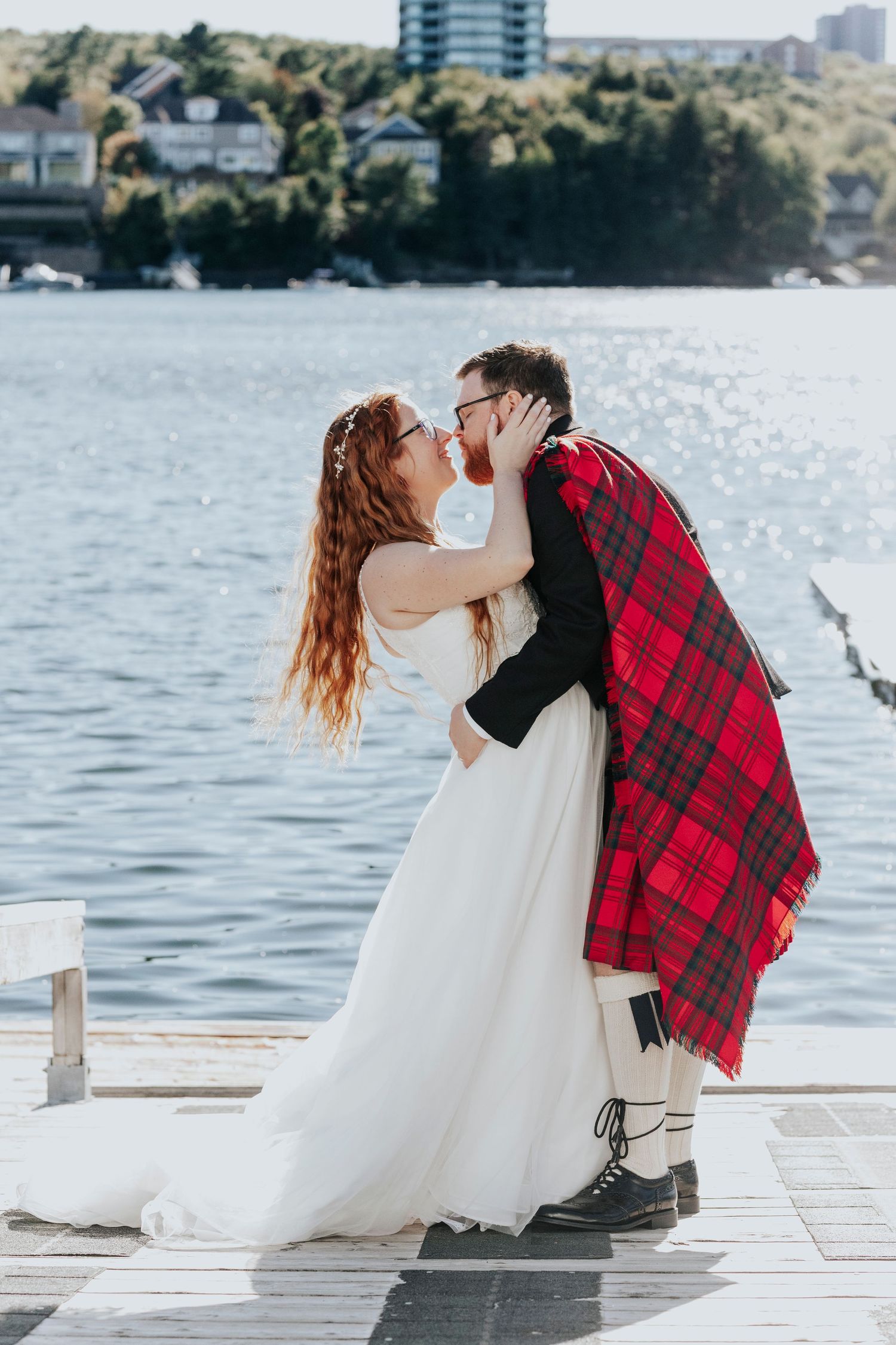 Bride and groom kiss on a sunny dock by the lake, groom wearing a red tartan kilt and traditional boots.