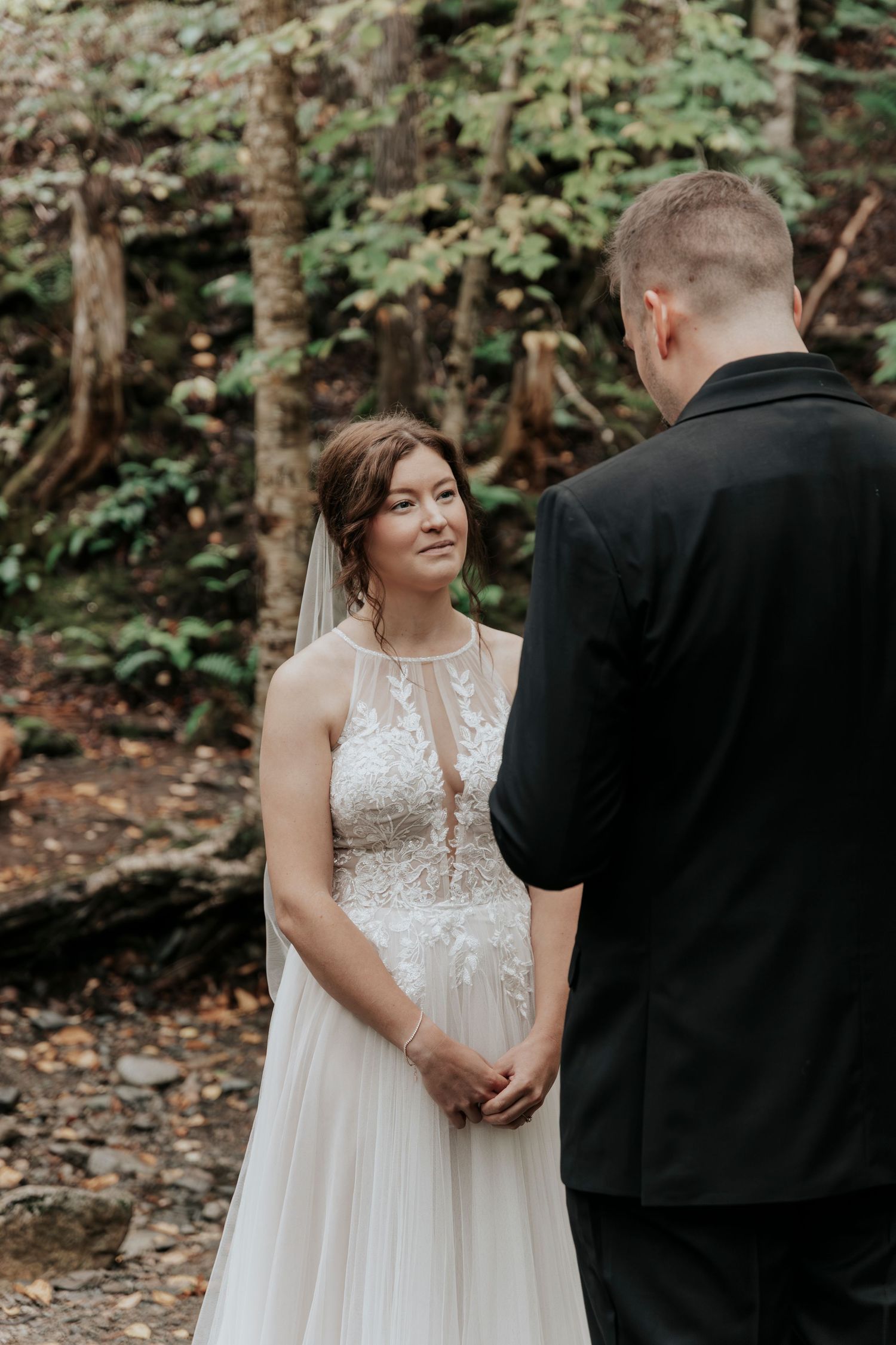 Bride gazes lovingly at her groom while holding hands during their woodland elopement vow exchange.