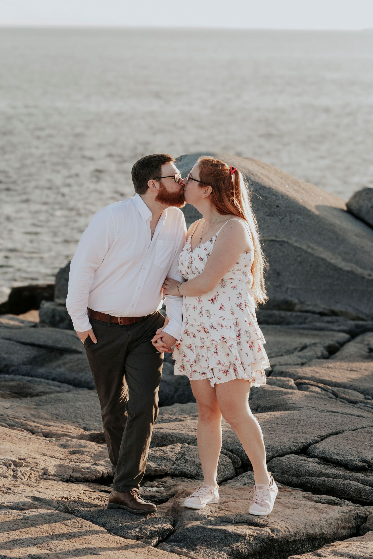 Couple sharing a kiss on rocky coastal shoreline with calm ocean waters and lighthouse in the background.