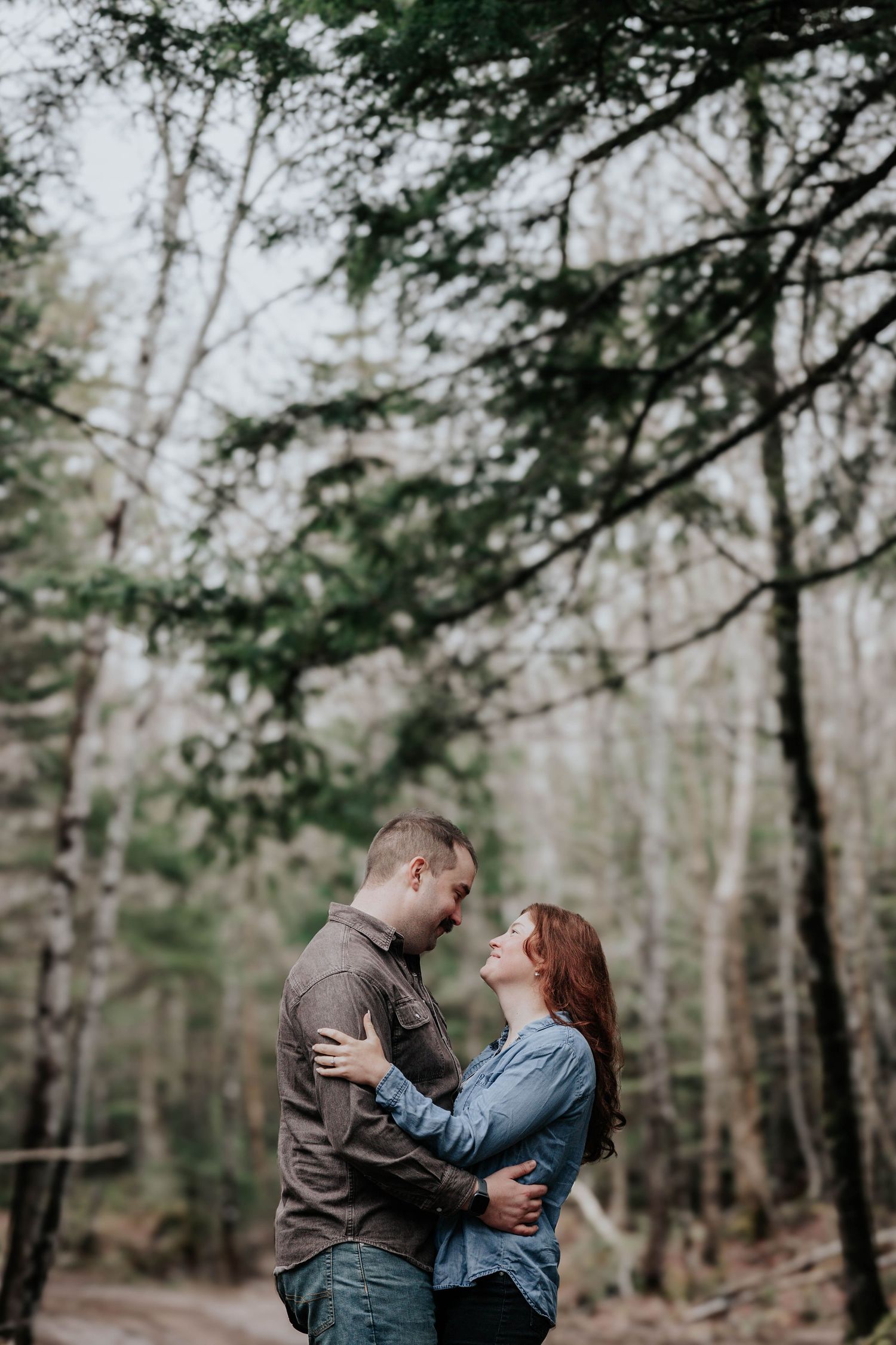 Couple embracing face to face on forest path surrounded by tall trees in soft light.